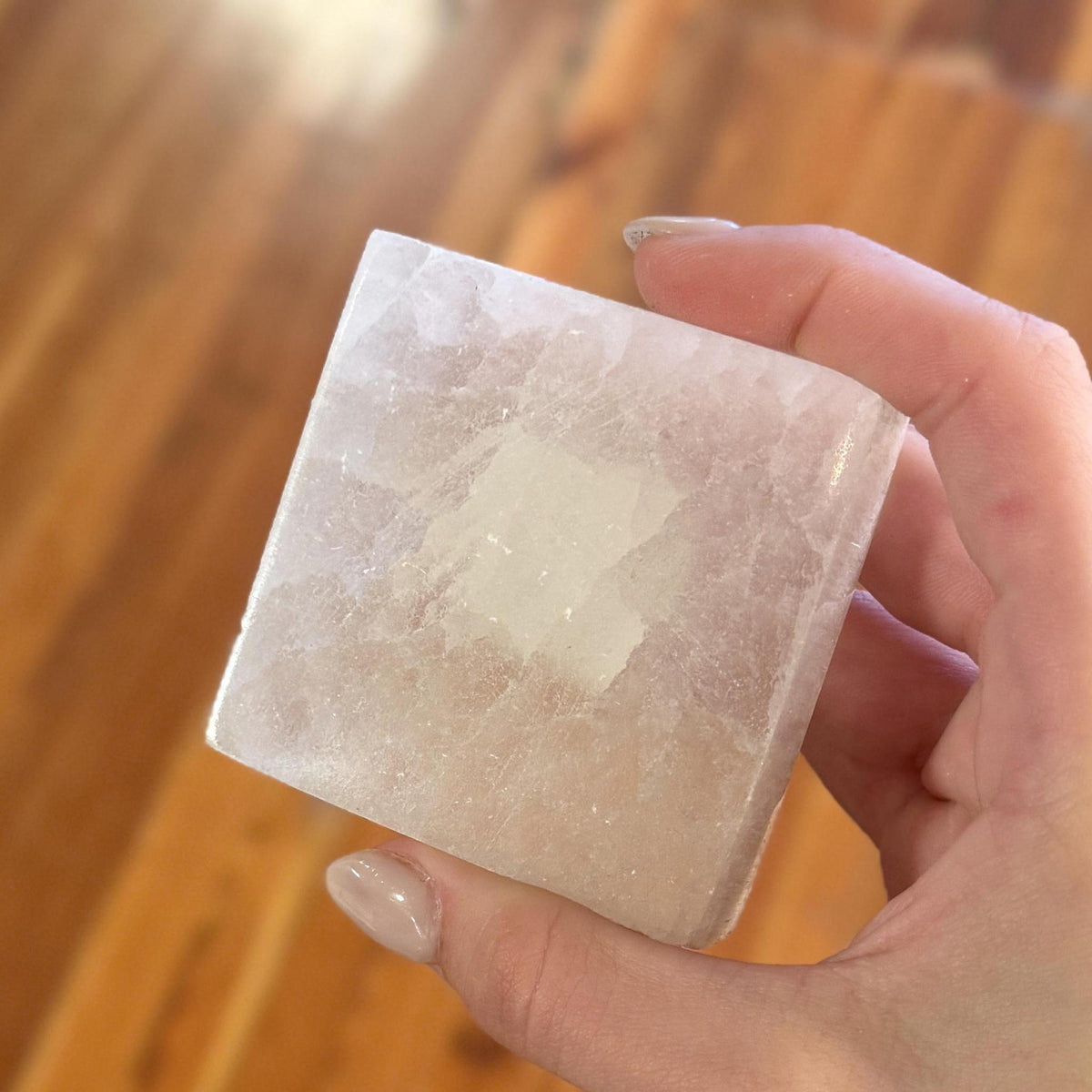 A hand with manicured nails holds the Spellcrafter Selenite Square Plate—a translucent white crystal—against a blurred wooden floor, radiating harmony.