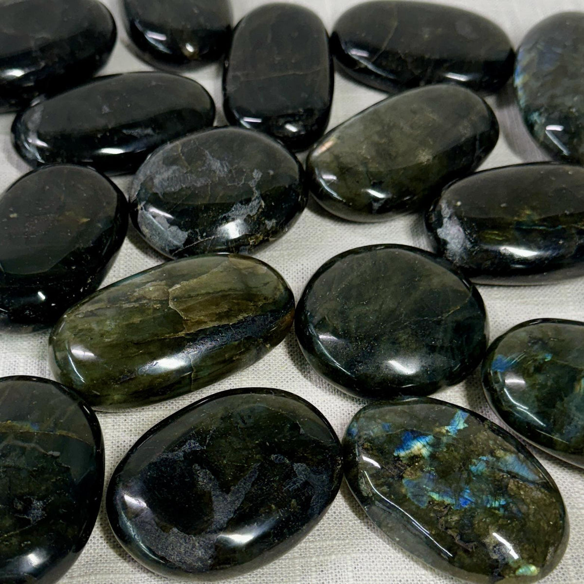 Spellcrafter's Labradorite Palm Stones feature polished, oval shapes with dark green, black, and iridescent blue hues—ideal for inspiring creativity and balancing chakras. Displayed on a light fabric background.