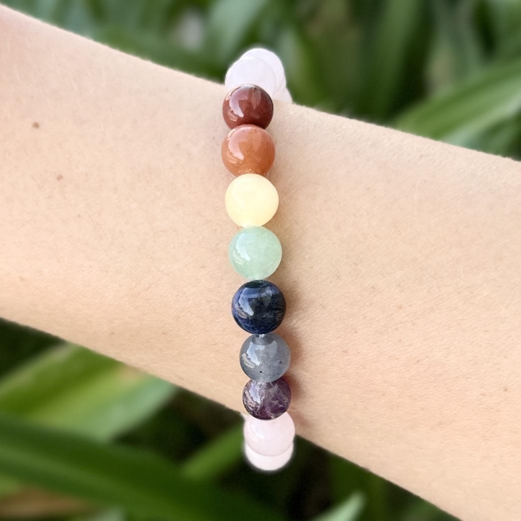 The Spellcrafter Chakra Stone Bracelet features colorful round beads in a gradient from brown, orange, yellow, green, blue, to purple and pink, shown on a wrist against a green leafy background.
