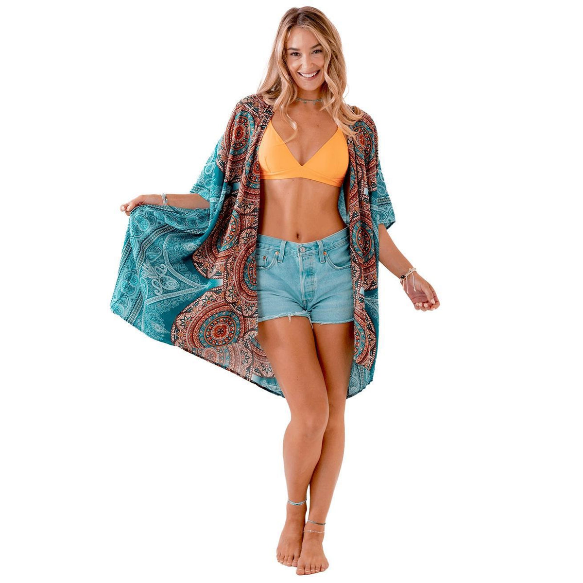 A woman poses barefoot against a white background, wearing the Spellcrafter Kimono - One Size over an orange bikini top and denim shorts, holding out the kimono's sides to display its colorful pattern.
