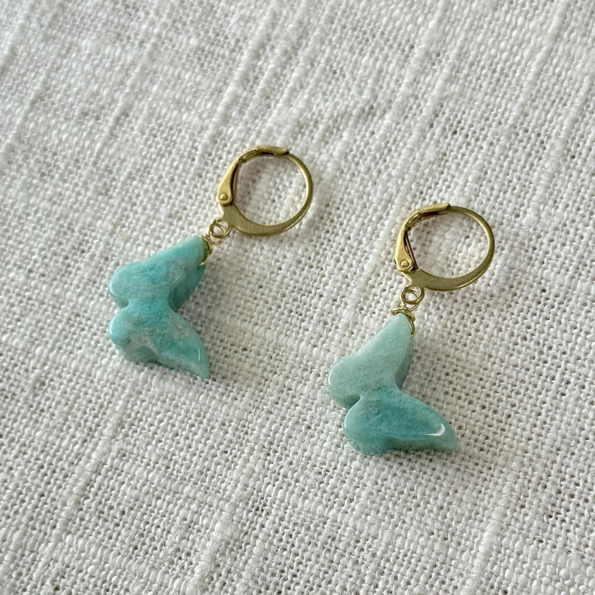 The Spellcrafter Amazonite Butterfly Earrings feature gold hoops adorned with light blue butterfly-shaped charms, shown on a textured white fabric background.