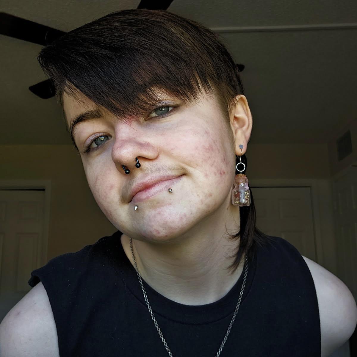 A person with short dark hair, facial piercings, and a sleeveless black top smiles...