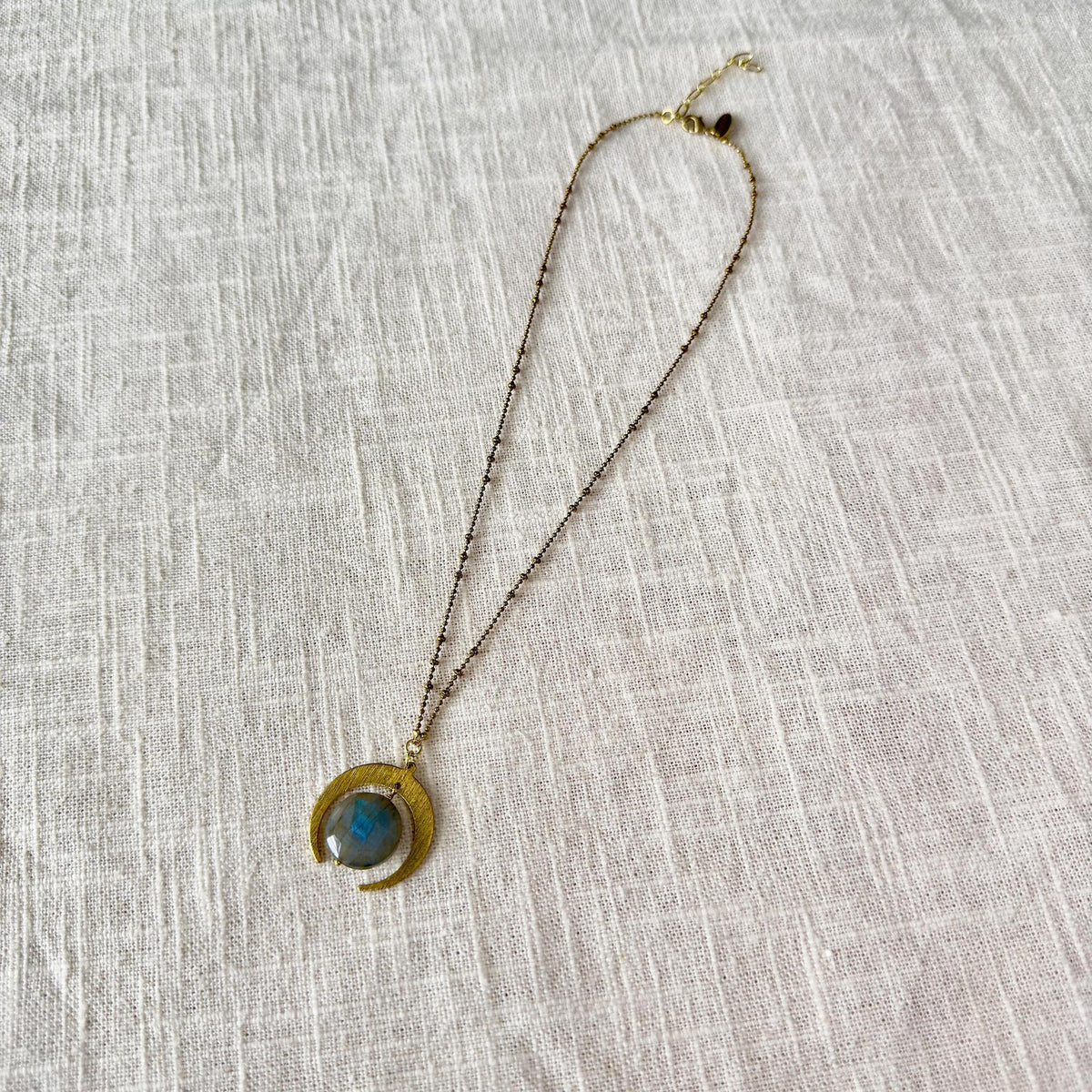 The Spellcrafter Labradorite Lunar Eclipse Necklace features a circular blue labradorite stone and crescent moon pendant, elegantly displayed against a textured beige fabric background.