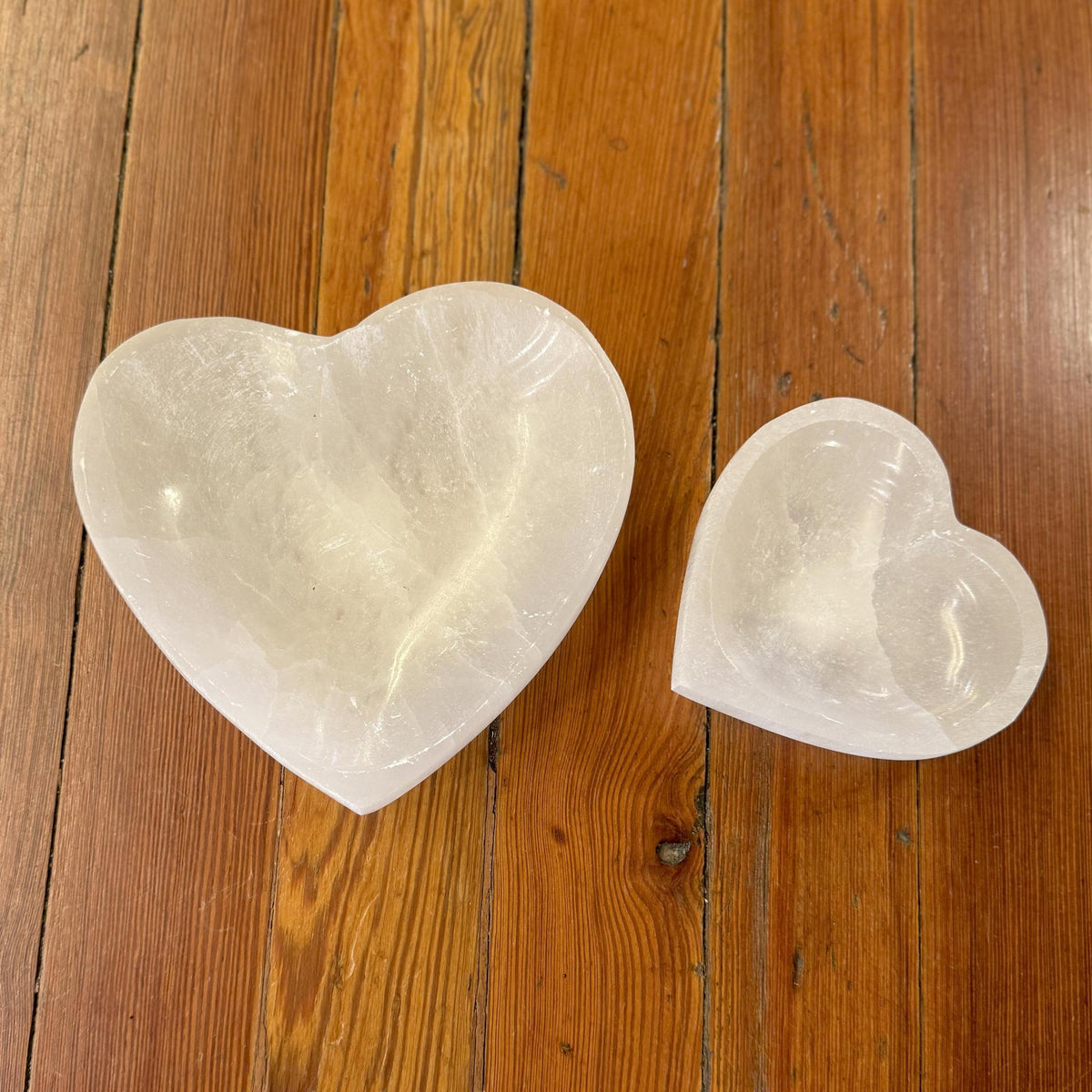 Two Spellcrafter Selenite Heart Shaped Bowls in different sizes rest on a wood floor, their white crystal surfaces bringing harmony and cleansing energy to any space.