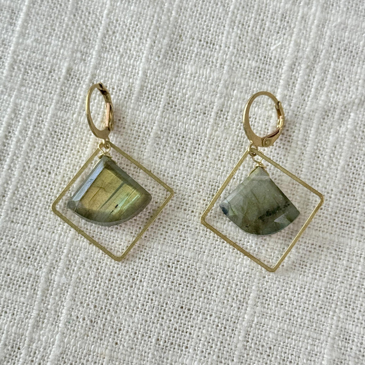 The Spellcrafter Labradorite Framed Diamond Earrings feature geometric square frames with translucent green labradorite stones, hypoallergenic brass construction, and are displayed on a textured white fabric background.
