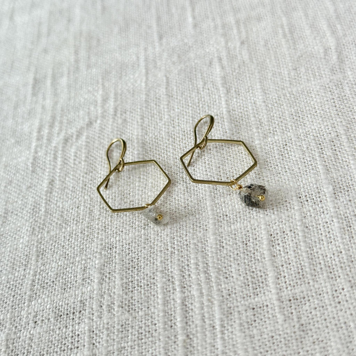 Spellcrafter’s Herkimer Hexagon Earrings feature gold hexagon hoops with dangling Herkimer Diamond meditation stones, shown on a textured white fabric background.