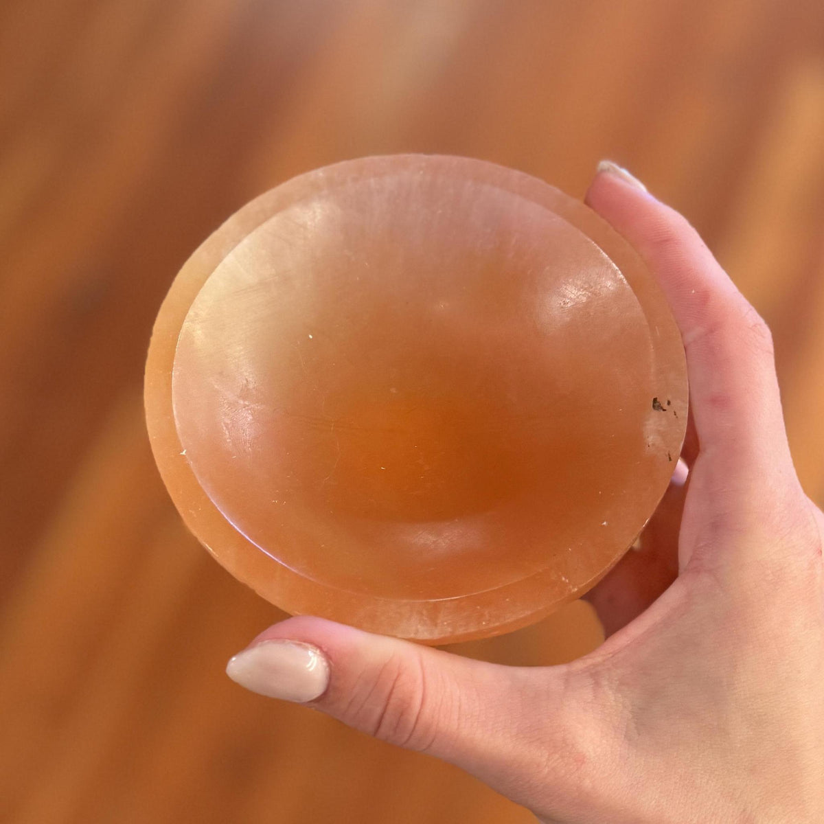 A manicured hand holds the Spellcrafter Medium Selenite Bowl—a round, pinkish-orange translucent dish ideal for cleansing crystals—set against a blurred wooden background.