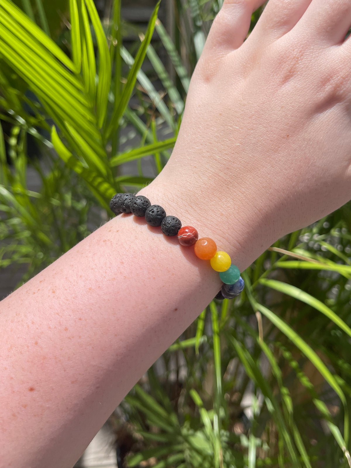 A hand wears the Spellcrafter Chakra Stone Bracelet, featuring black stones and vibrant beads, set against a lush green plant backdrop.