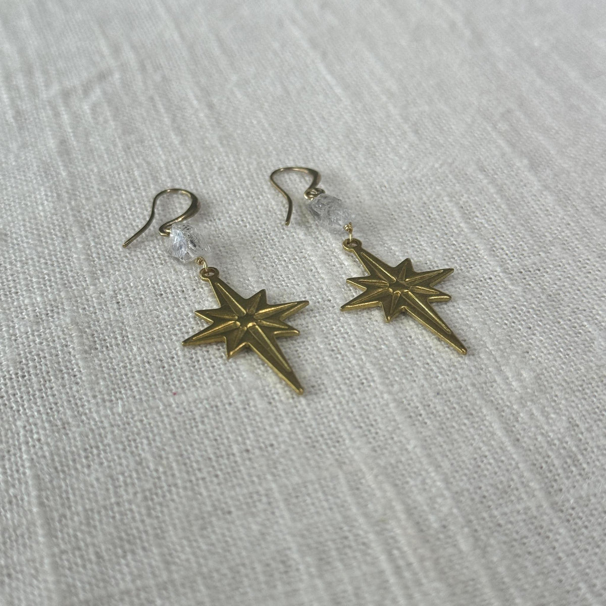 The Spellcrafter Herkimer Diamond North Star Earring features gold star-shaped dangles with clear beads, inspired by Herkimer Diamond energy, displayed on a textured white fabric background.
