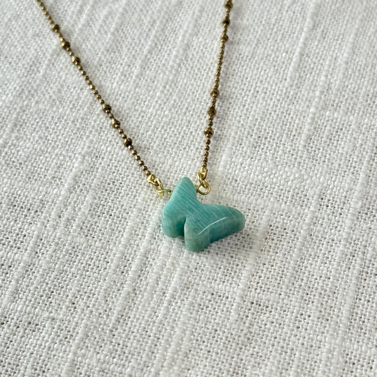 The Spellcrafter Amazonite Butterfly Necklace features a gold beaded chain and a light blue amazonite butterfly pendant, known for chakra balancing, displayed on a textured off-white fabric background.