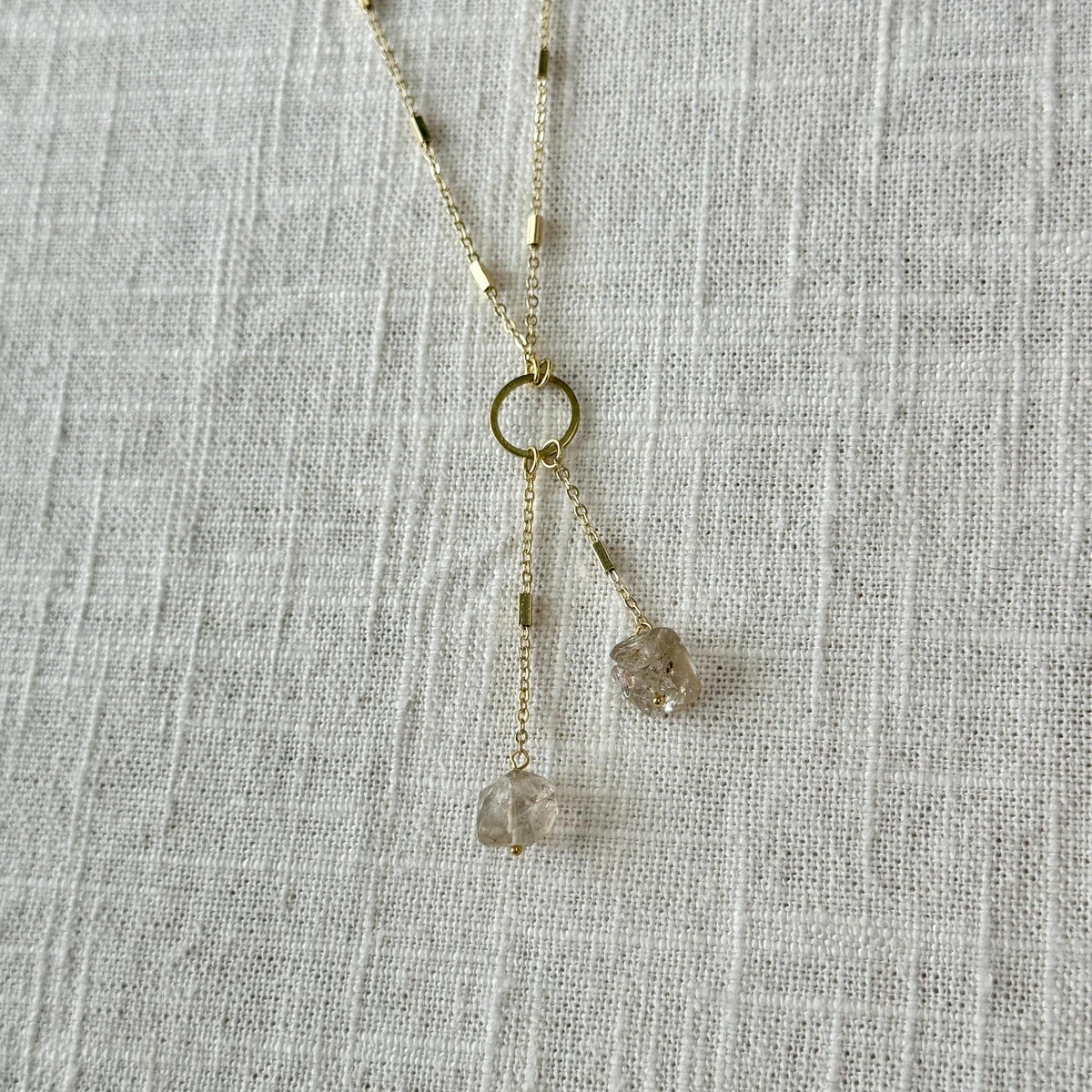 The Spellcrafter Herkimer Diamond Tassel is a gold necklace with two thin chains and rough Herkimer Diamonds, perfect for meditation or Crown chakra activation, shown against a textured white fabric background.
