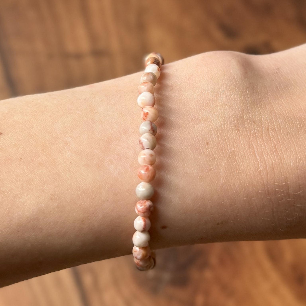 A close-up of a wrist wearing the Spellcrafter Red Vein Jasper Crystal Bracelet, featuring small light pink, white marbled, and Red Vein Jasper beads—revered as a recovery stone for the Root Chakra—with a blurred wooden background.