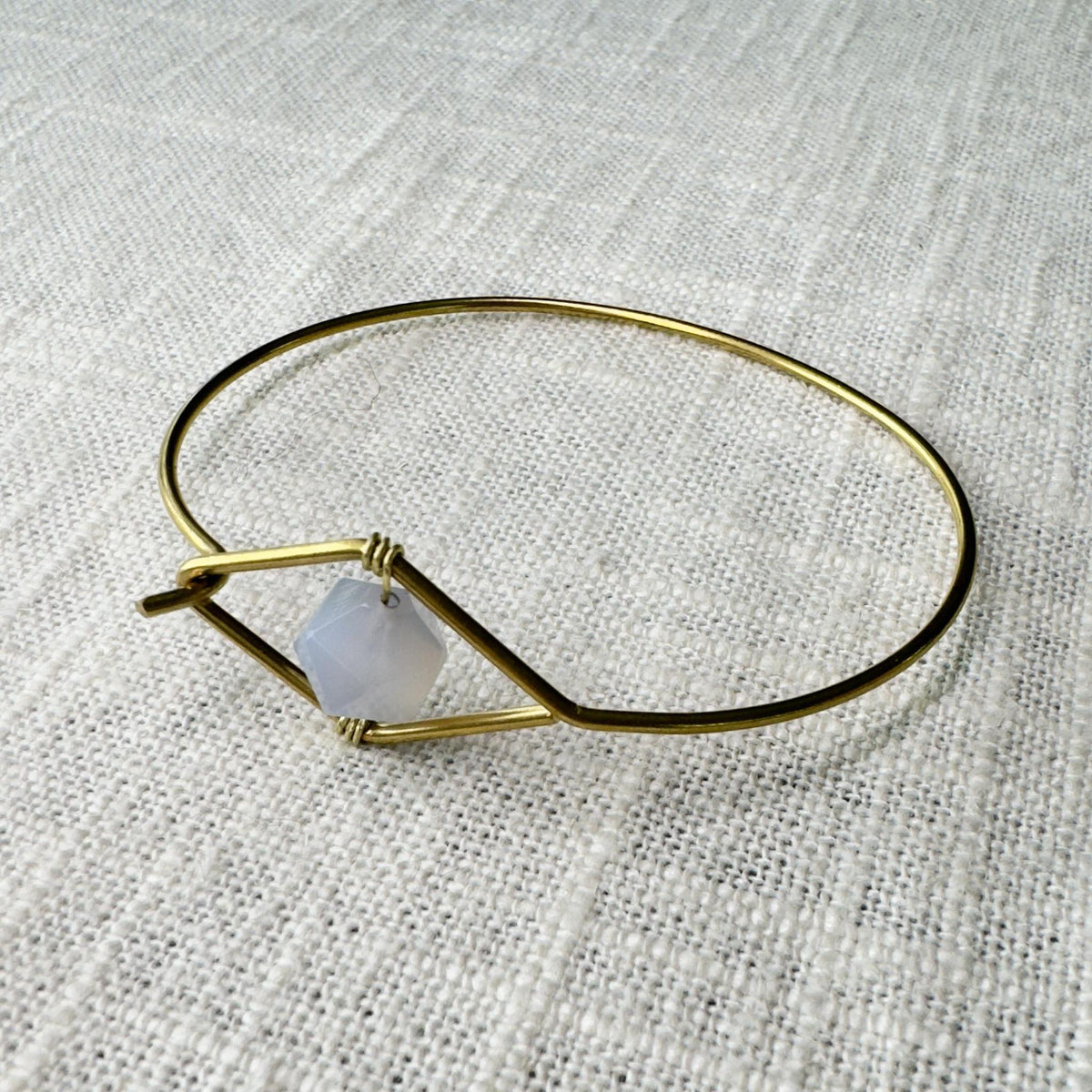 The Spellcrafter Blue Lace Agate Wire Bracelet features a faceted gemstone symbolizing the Throat chakra, set in a geometric gold design and showcased on textured beige fabric.
