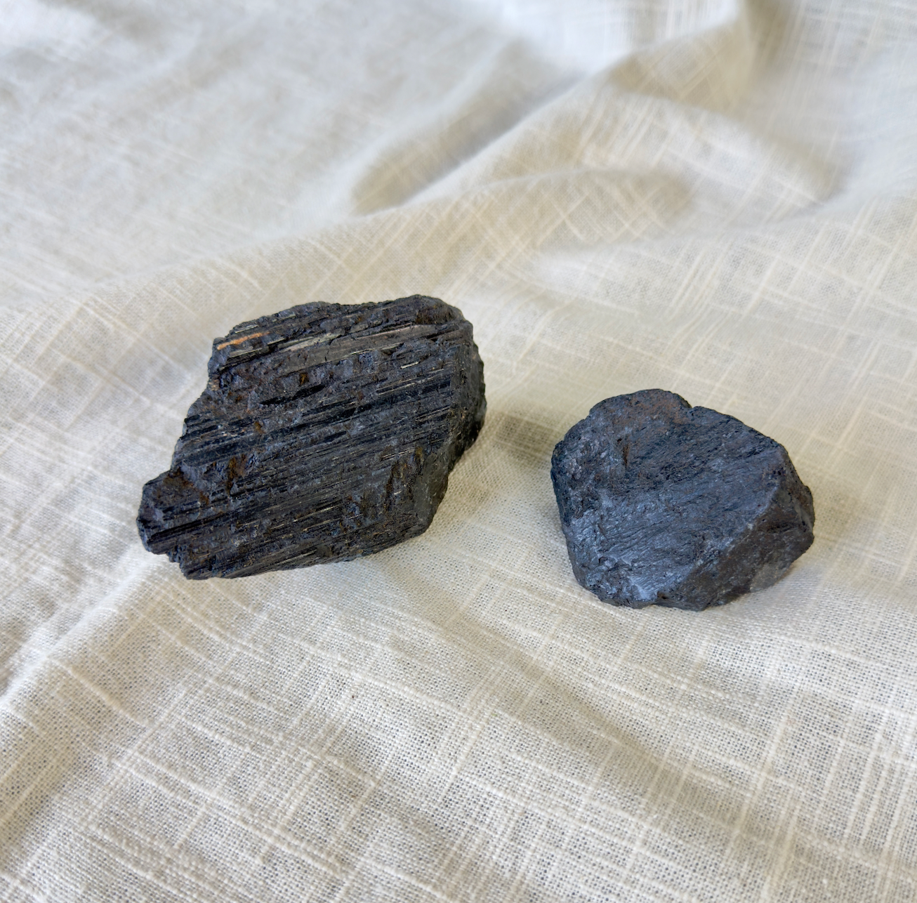 Two dark, rough Spellcrafter Black Tourmaline Rough Chunks rest on textured fabric. The left...