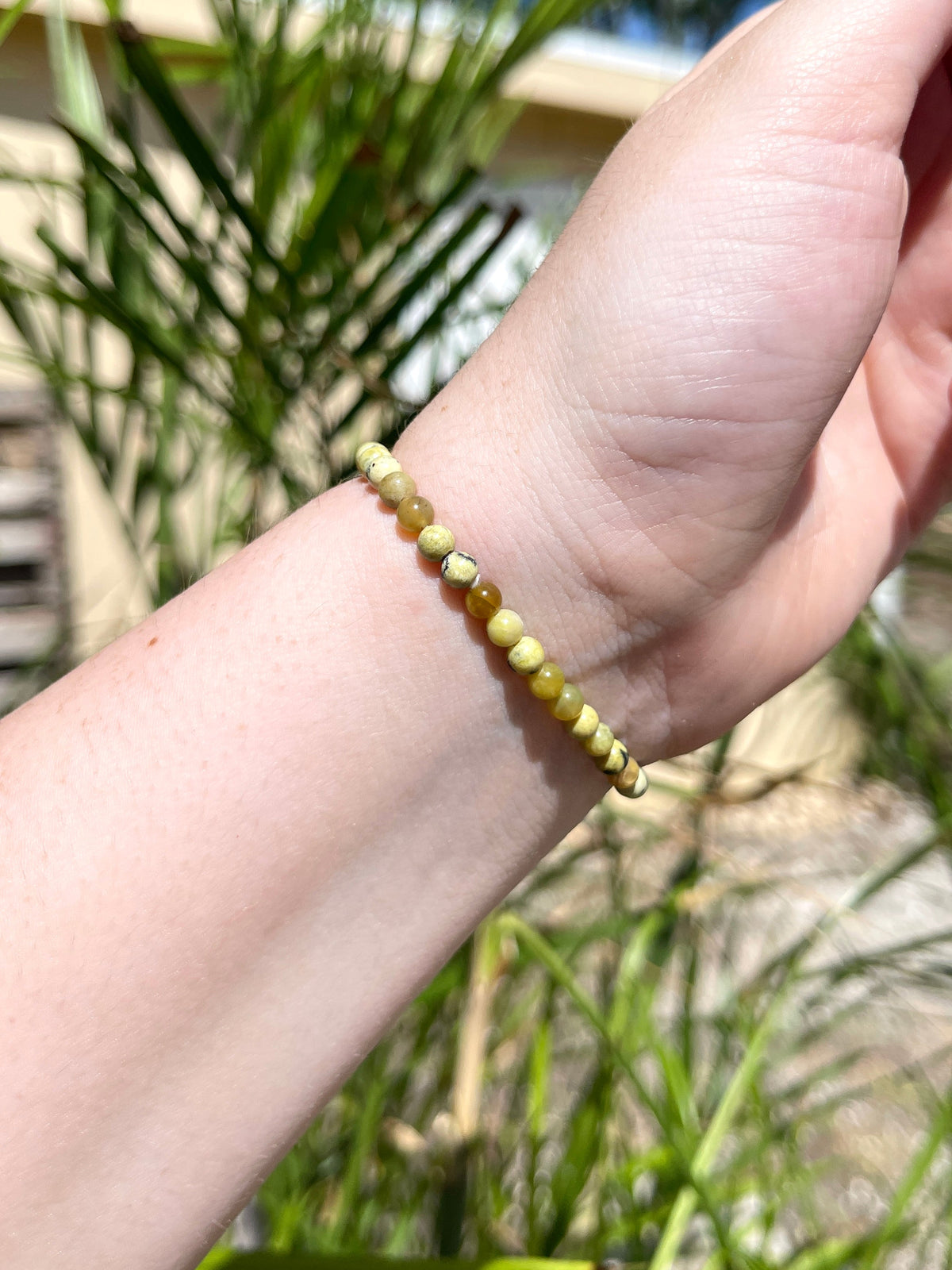 A hand models the Spellcrafter Yellow Agate Bracelet, featuring small round yellow-green beads, outdoors with greenery and a blurred building in the background.