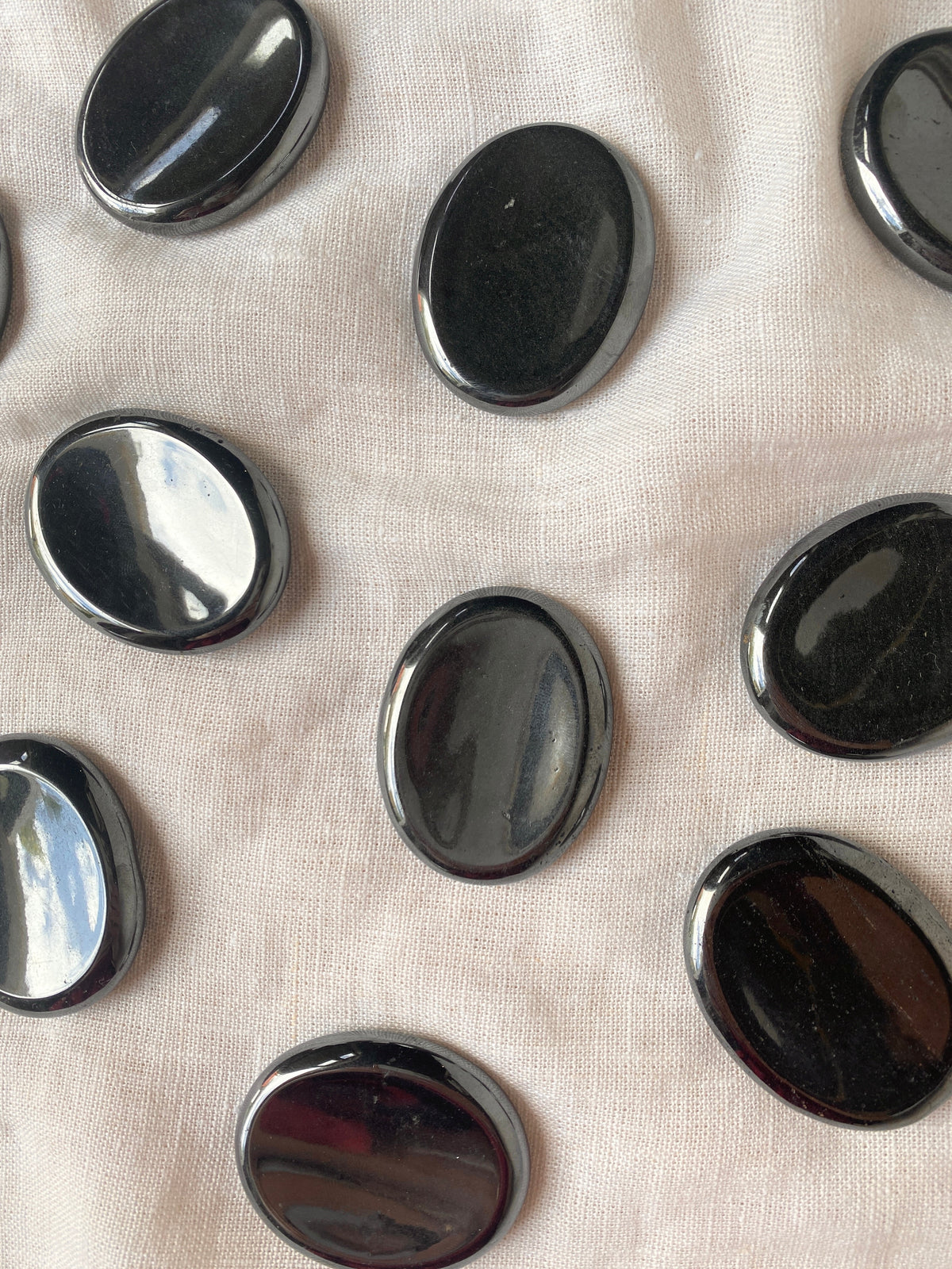 Spellcrafter Hematite Calming Stones, smooth and oval-shaped, are arranged on light beige fabric with some stones reflecting light.