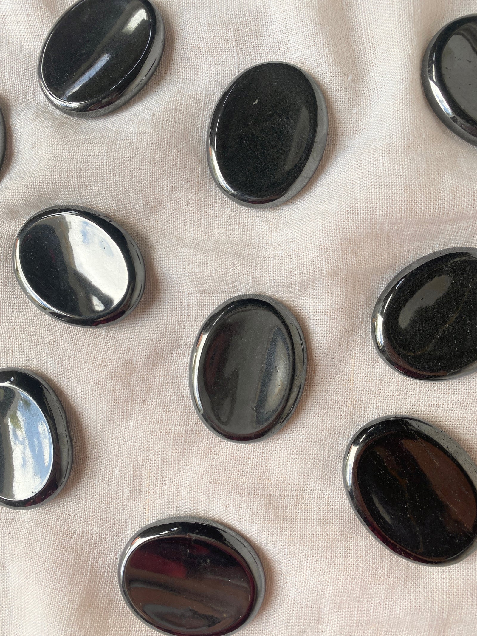 Spellcrafter Hematite Calming Stones, smooth and oval-shaped, are arranged on light beige fabric with...