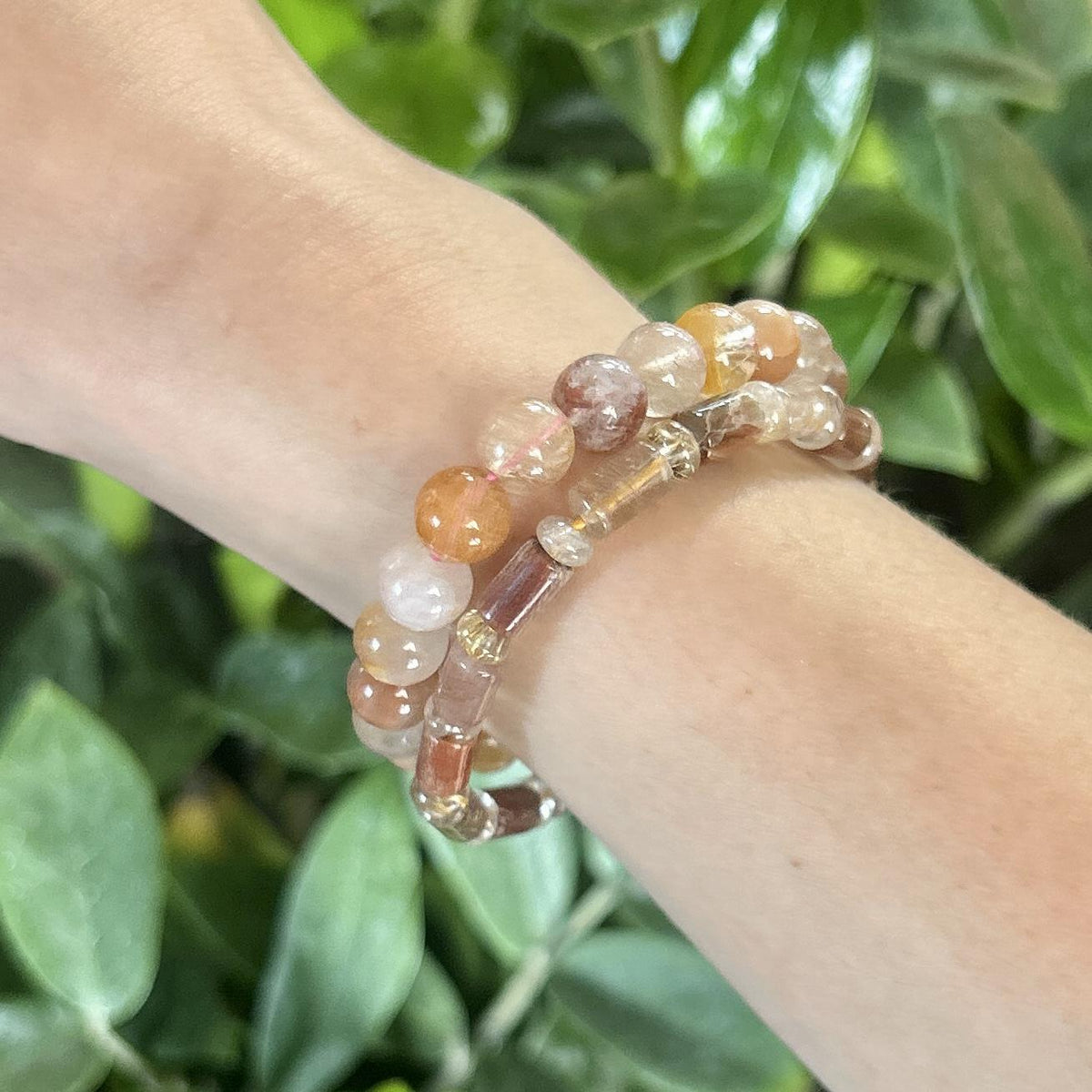 A wrist adorned with two Spellcrafter Golden Healer Crystal Bracelets, their polished, multicolored beads shining brightly against a lush green leaf backdrop.