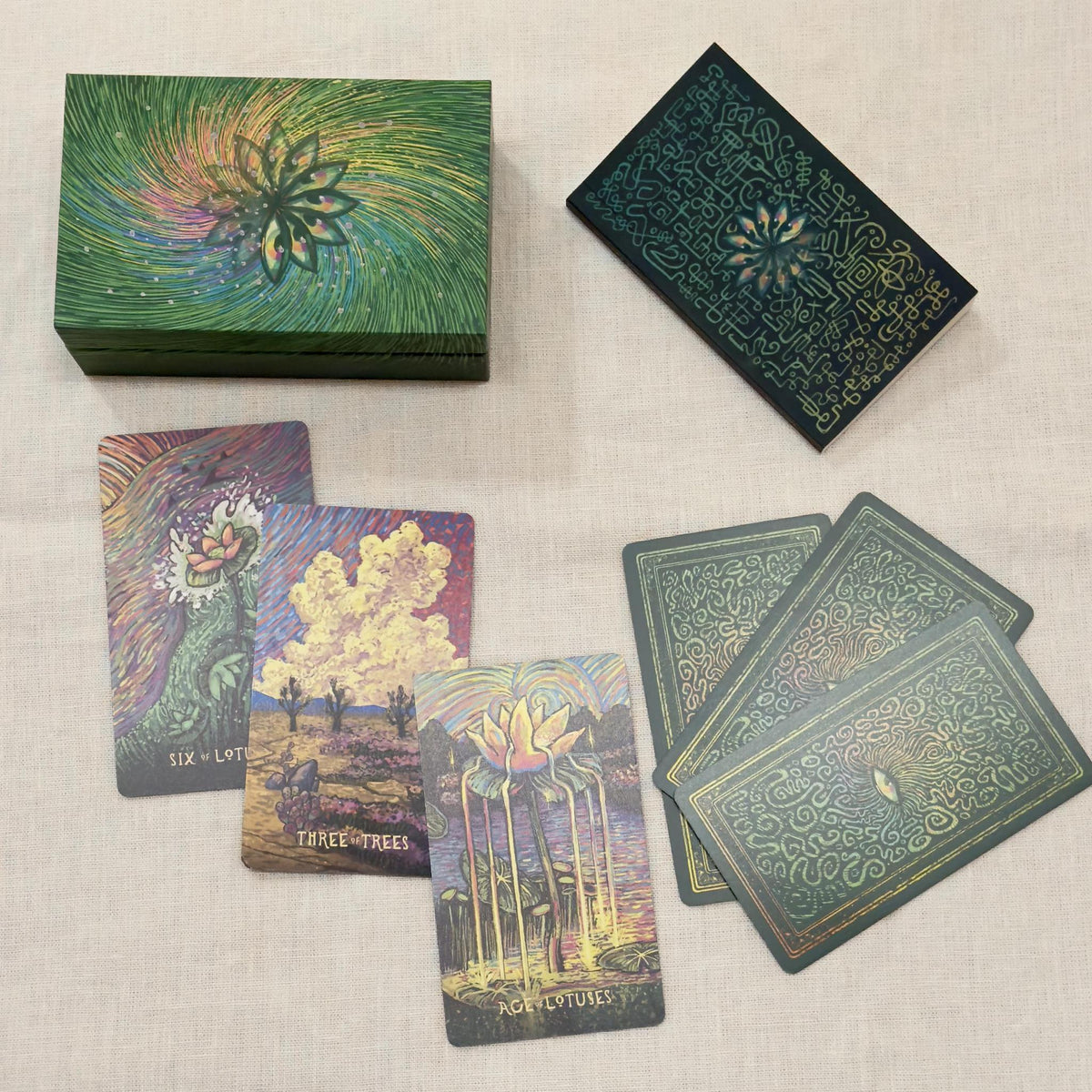 A Spellcrafter Cosma Visions Oracle deck with illustrated cards rests on a beige surface. Three face-up cards depict a lotus, trees, and clouds, while others lie face down next to a decorative green and gold box.