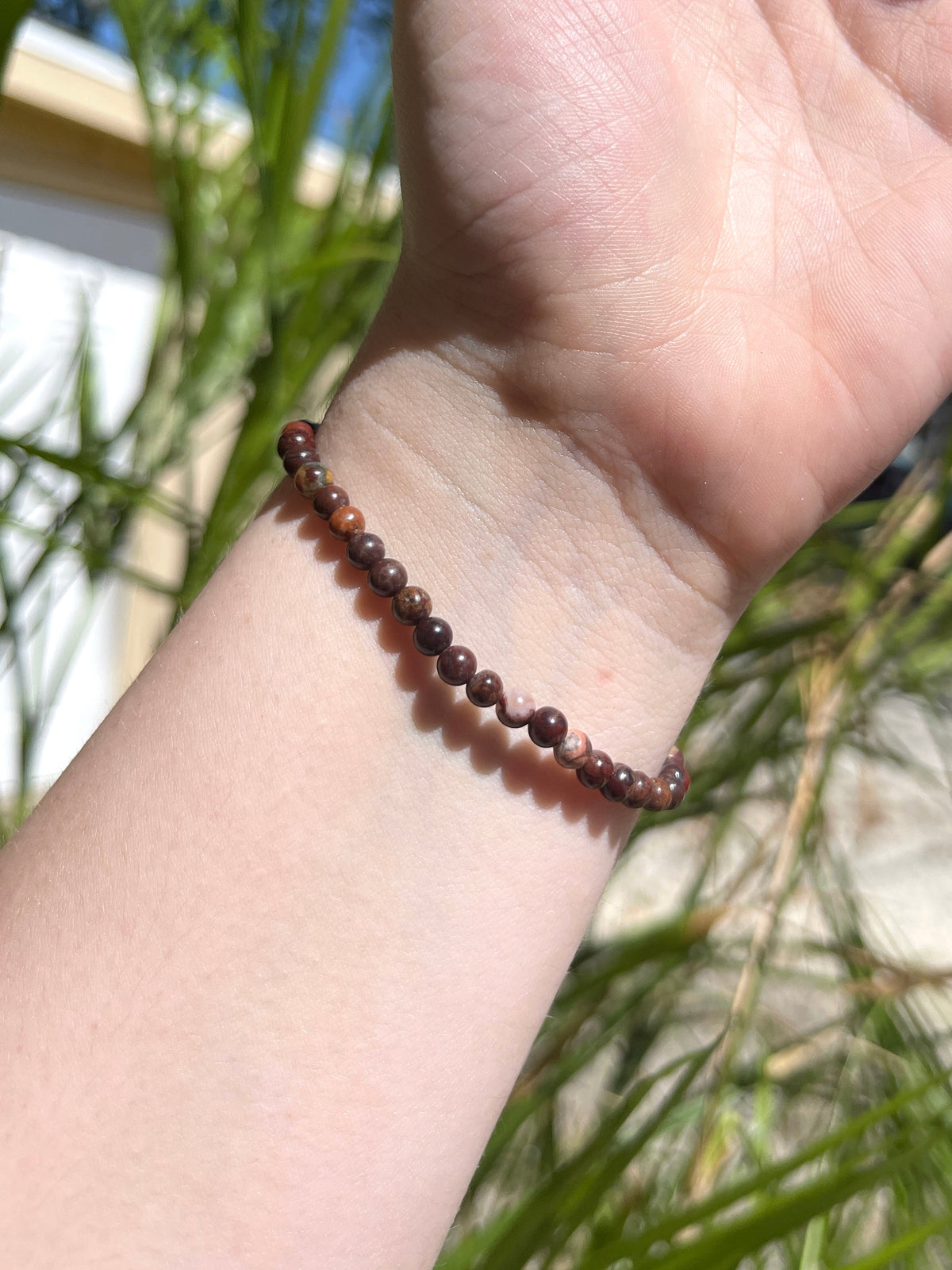 A close-up of a wrist adorned with Spellcrafter's Jasper Bracelet—thin, with small round brown beads—sunlit skin and blurred green plants in the background.