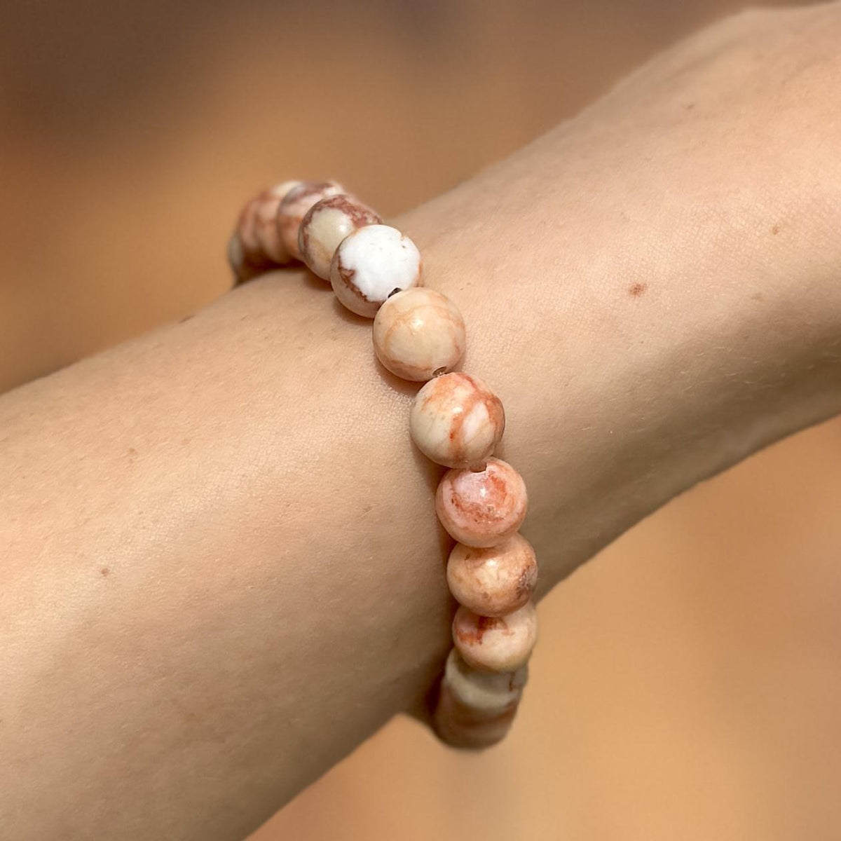 A close-up of a wrist wearing the Spellcrafter Red Vein Jasper Crystal Bracelet, crafted with marbled white, cream, and light brown beads—Red Vein Jasper is a powerful Root Chakra recovery stone. The warm-toned background is softly blurred.