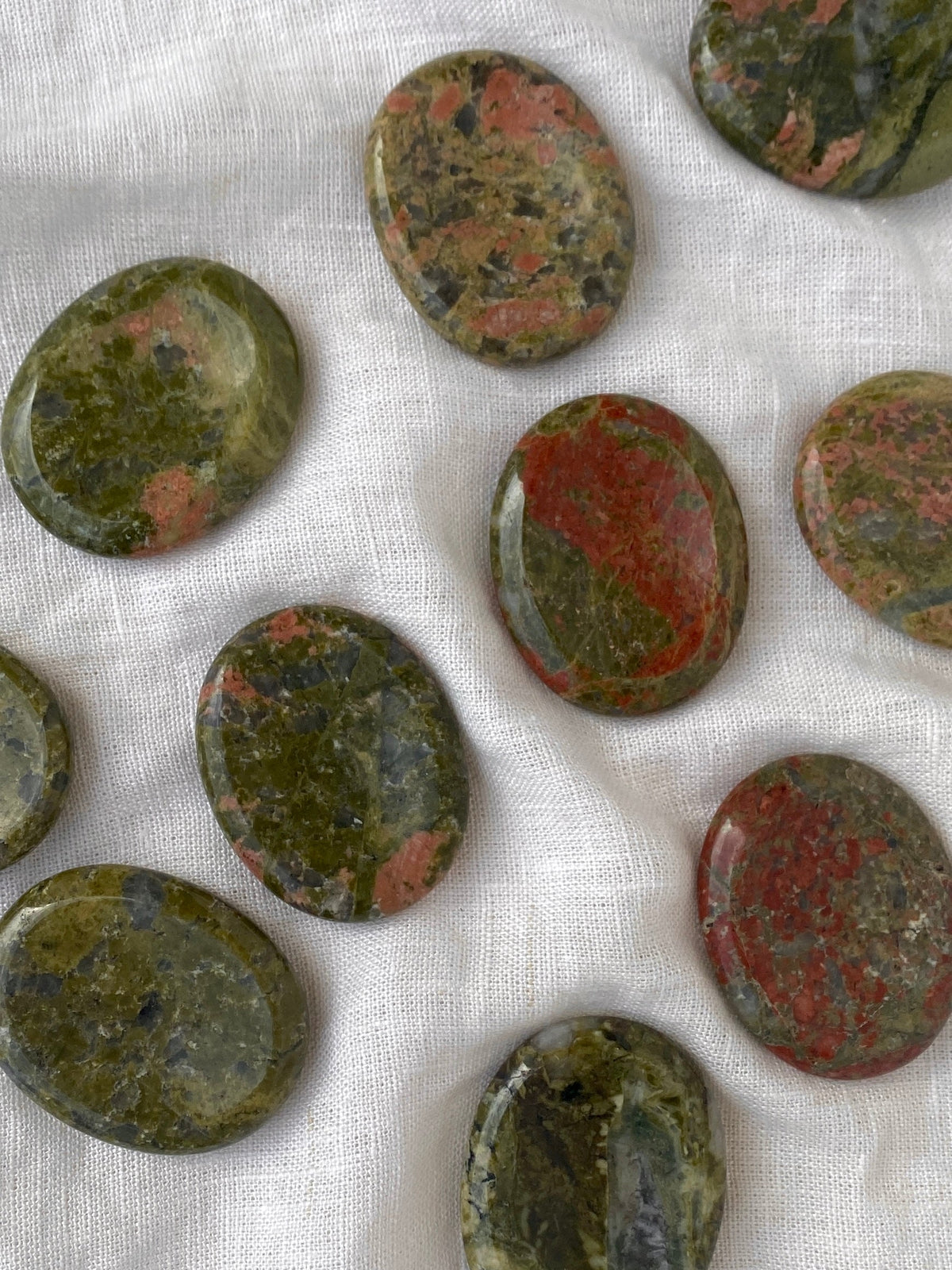 Oval, polished Spellcrafter Unakite Calming stones featuring green and reddish-pink patterns are displayed on a white textured fabric background.