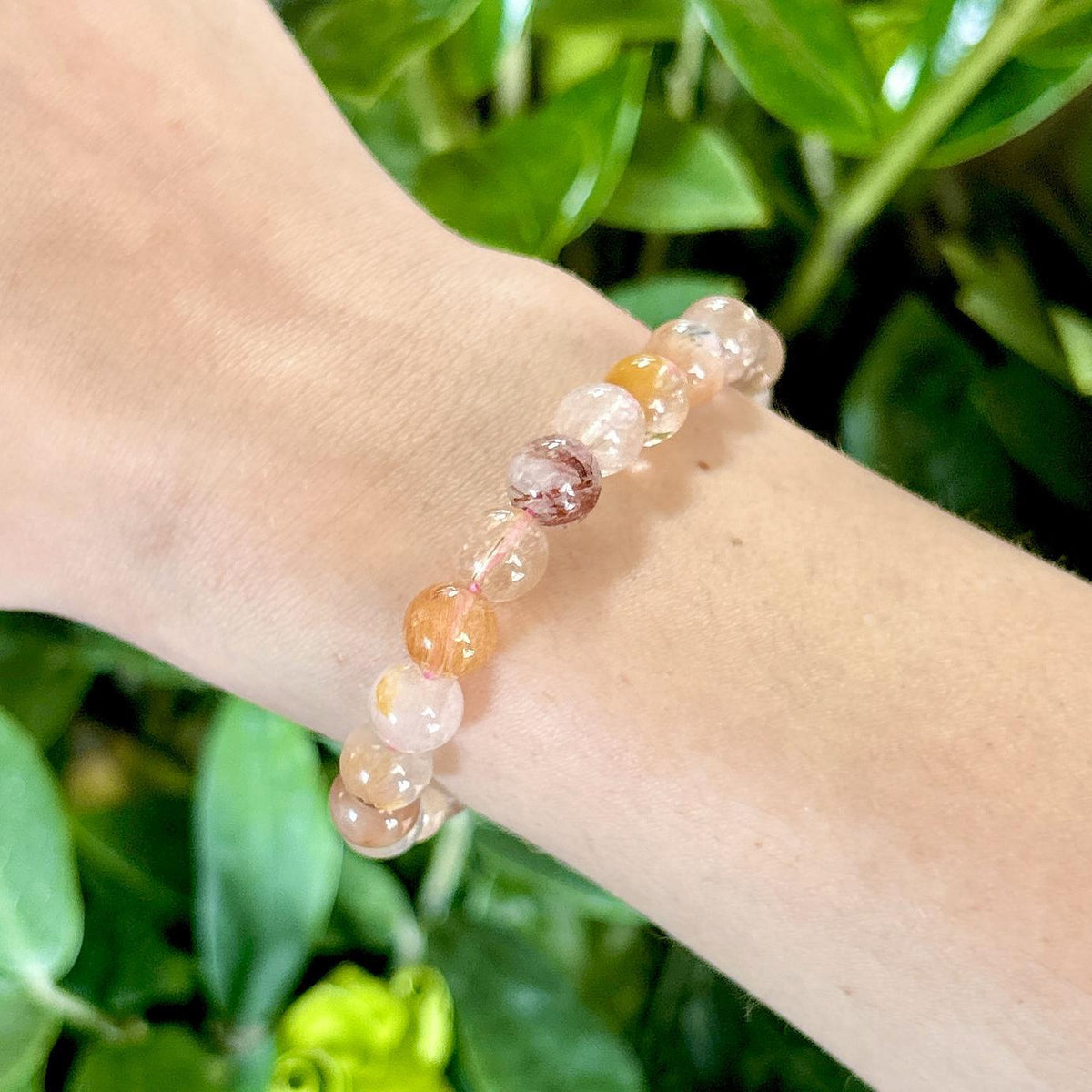 A light-skinned wrist wears the Spellcrafter Golden Healer Crystal Bracelet, featuring round translucent beads in pink, yellow, and orange tones. Green leafy plants appear in the background.