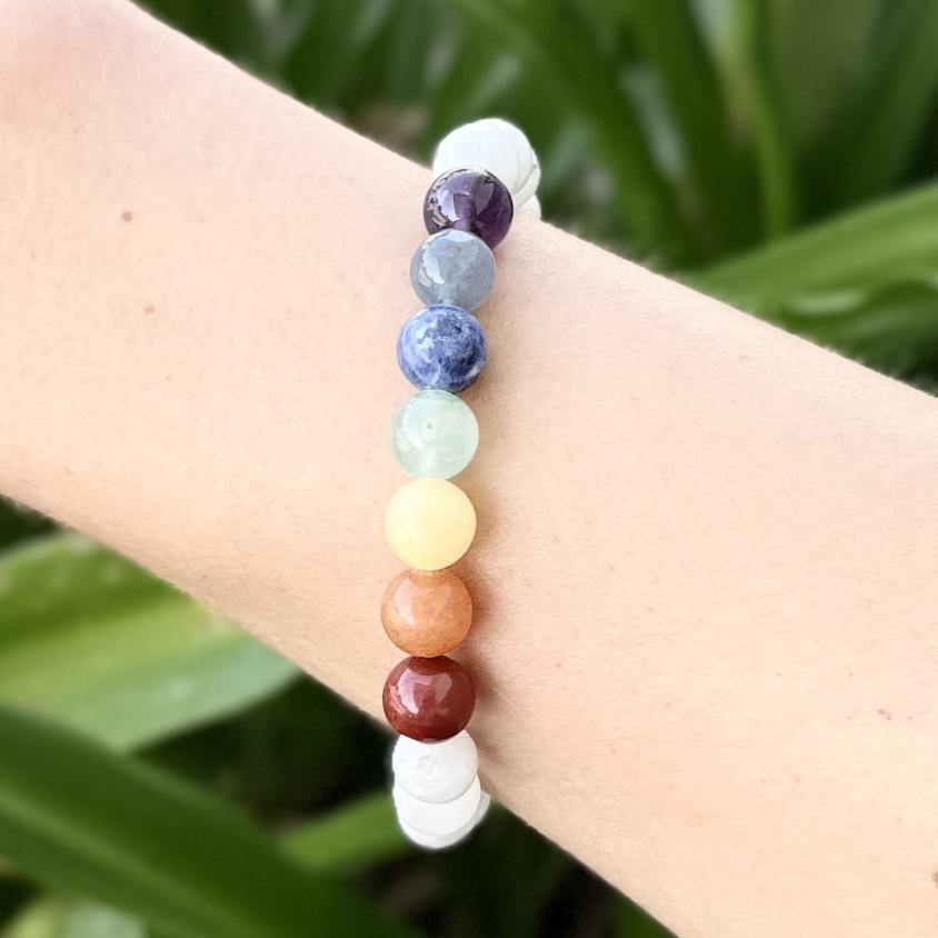 A close-up shows a person's wrist wearing the Spellcrafter Chakra Stone Bracelet, featuring seven different colorful stones in a row, set against a blurred green outdoor background.