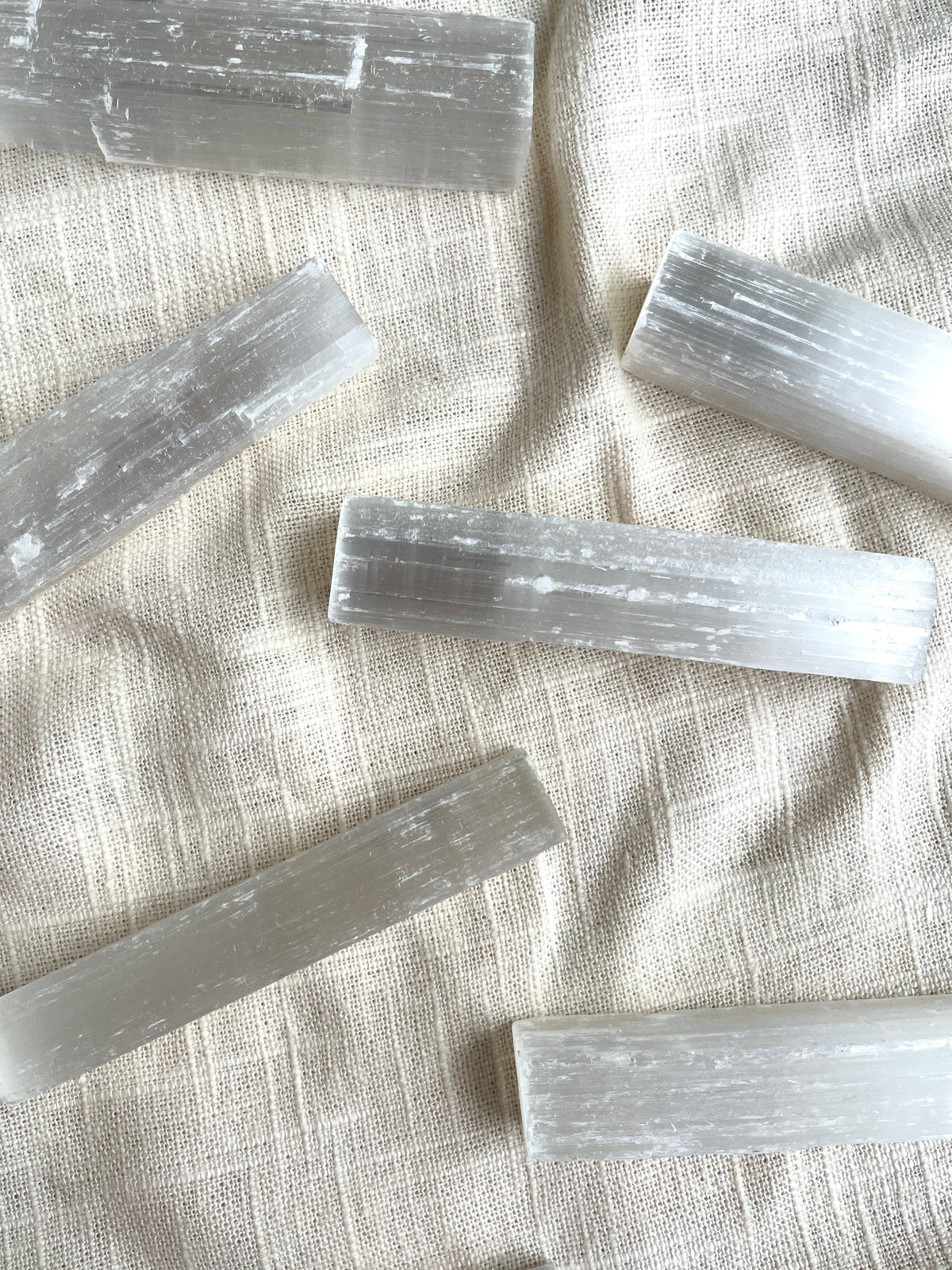 Spellcrafter Selenite Sticks rest on a textured off-white fabric, their smooth, striated surfaces glowing in soft natural light—ideal for cleansing and dispelling negative energy.