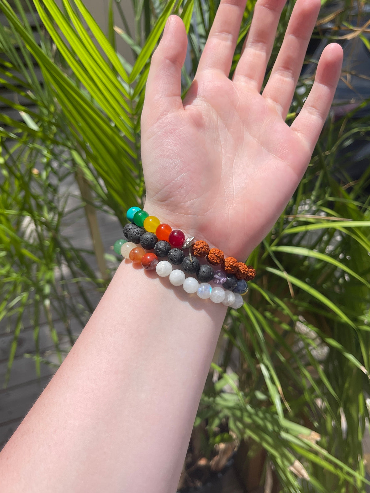 A hand is held up outdoors in sunlight, wearing three Spellcrafter Chakra Stone Bracelets with colorful stones and natural beads. Green palm leaves appear softly blurred in the background.