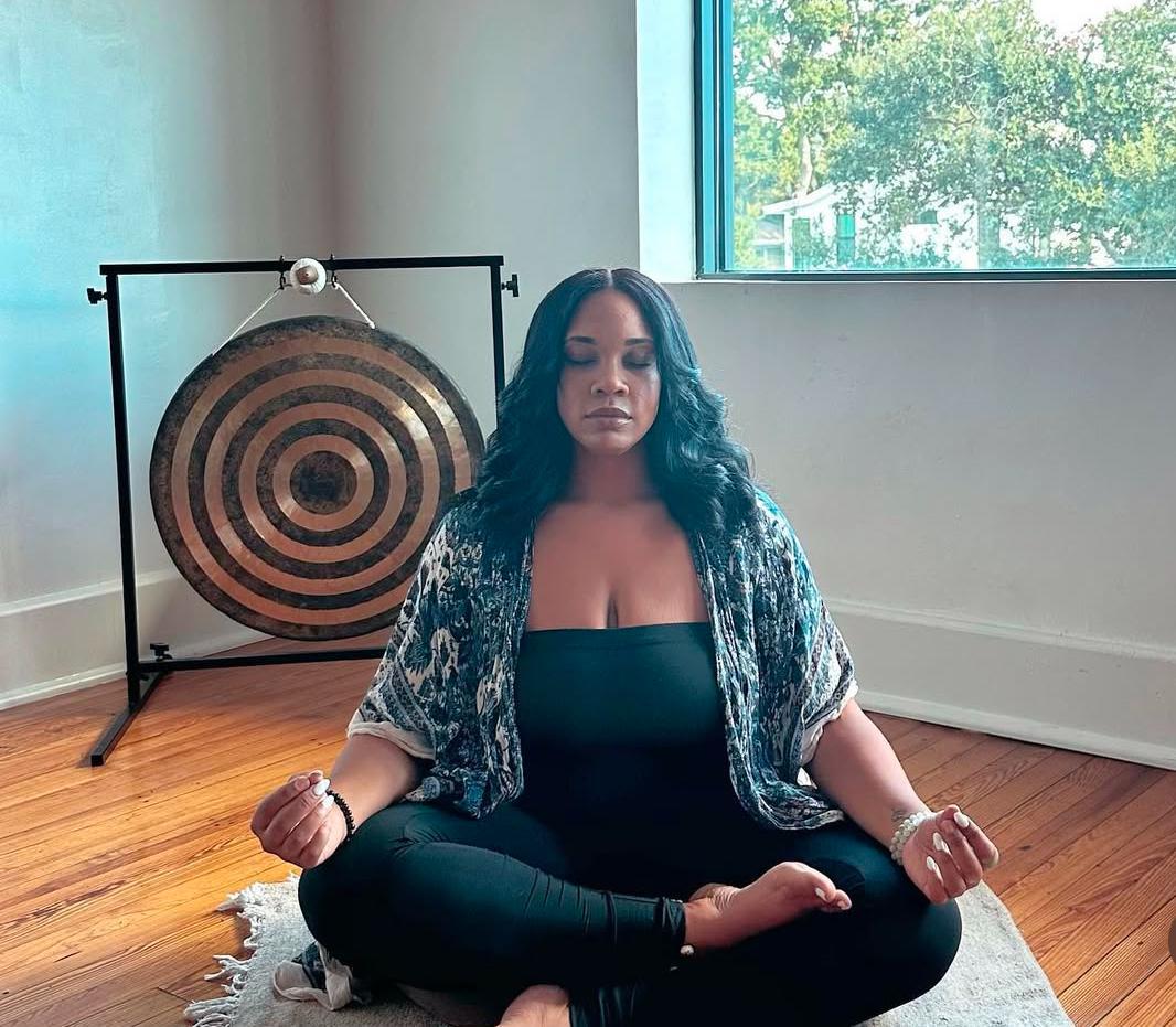 A woman sits cross-legged indoors, meditating in front of a large gong, as sunlight...