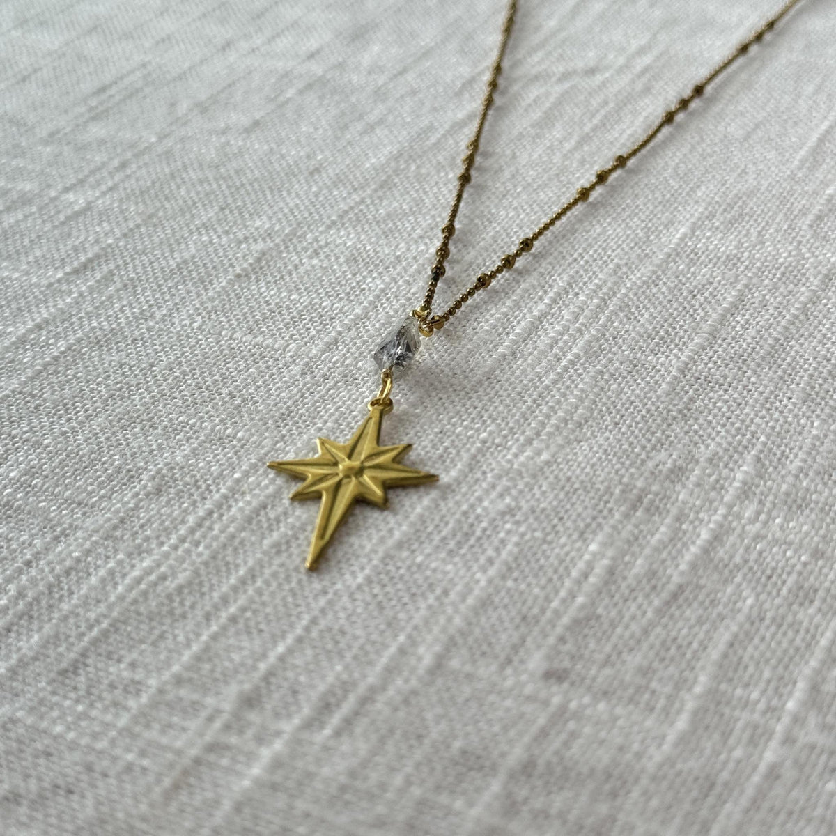 The Spellcrafter Herkimer Diamond North Star Necklace, featuring an eight-pointed star pendant and sparkling Herkimer Diamond, rests on a textured, light-colored fabric background.