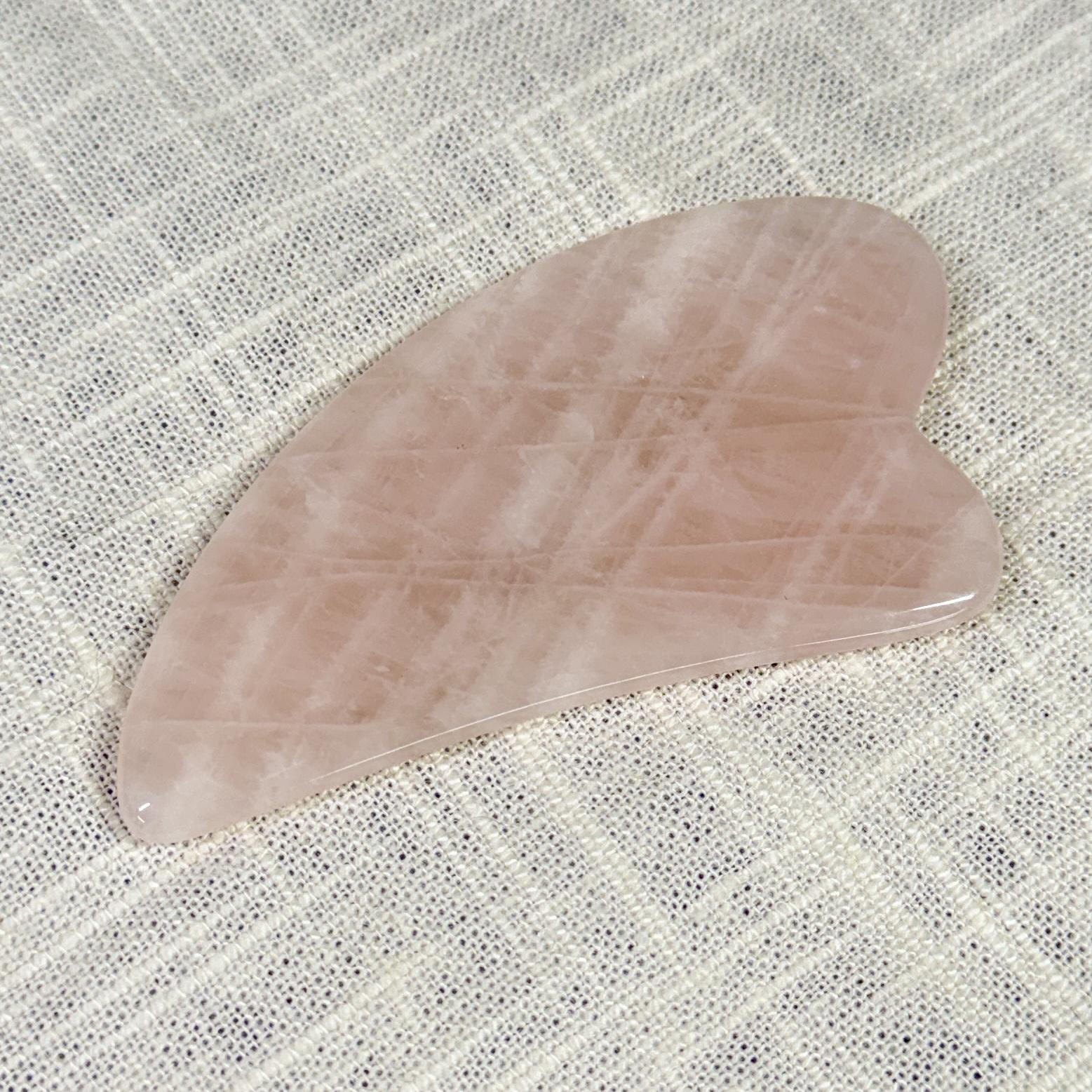 The Spellcrafter Rose Quartz Gua Sha, featuring smooth, curved edges, rests on a textured,...