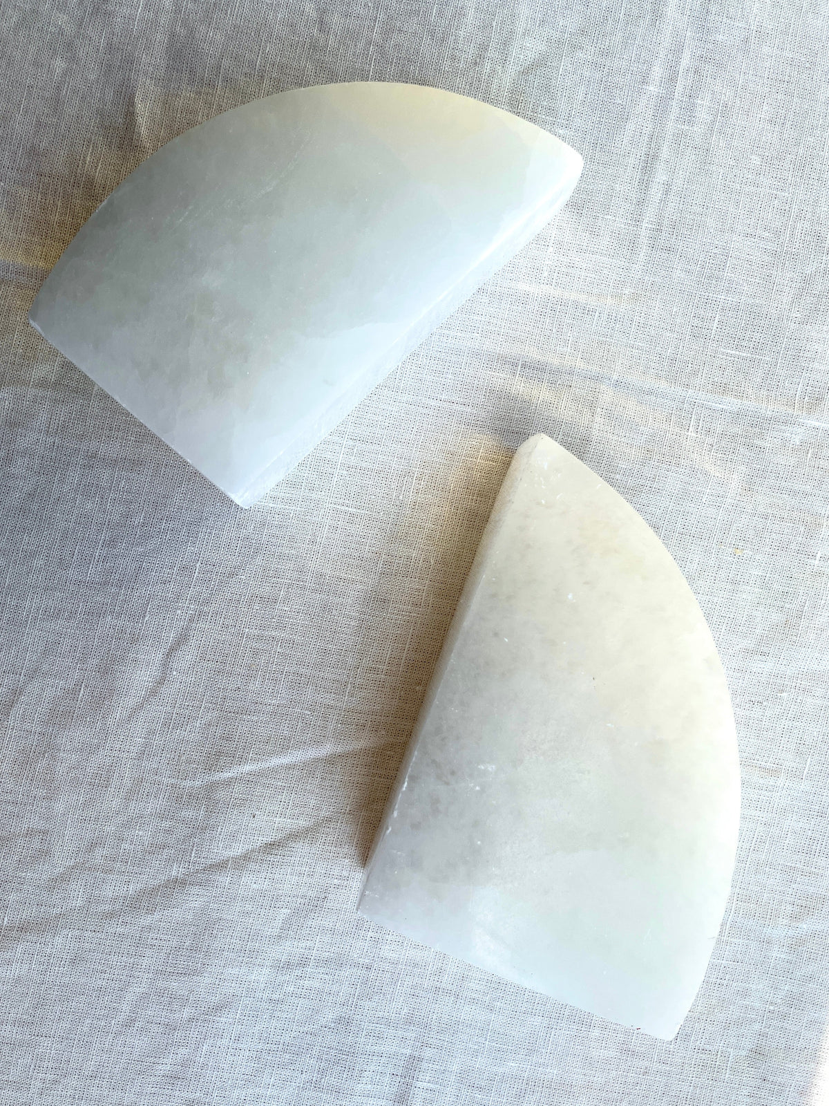 Two white, semi-circular Spellcrafter Selenite Bookends rest on a light, textured fabric. Polished and elegantly arranged at different angles, they add symmetry and style to your home décor.