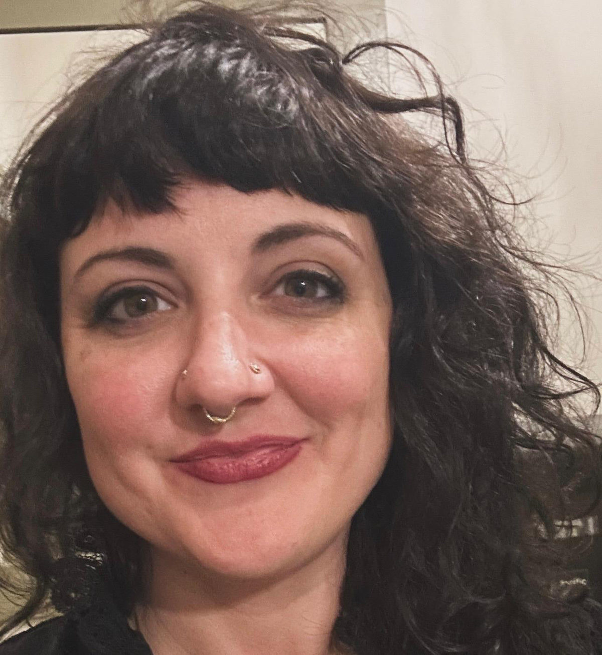 A woman with wavy dark hair and bold makeup smiles at the camera, embodying Spellcrafter’s mystical energy. Her look sets the tone for an in-person "Reading with Jess" tarot session—the background is neutral and softly blurred.