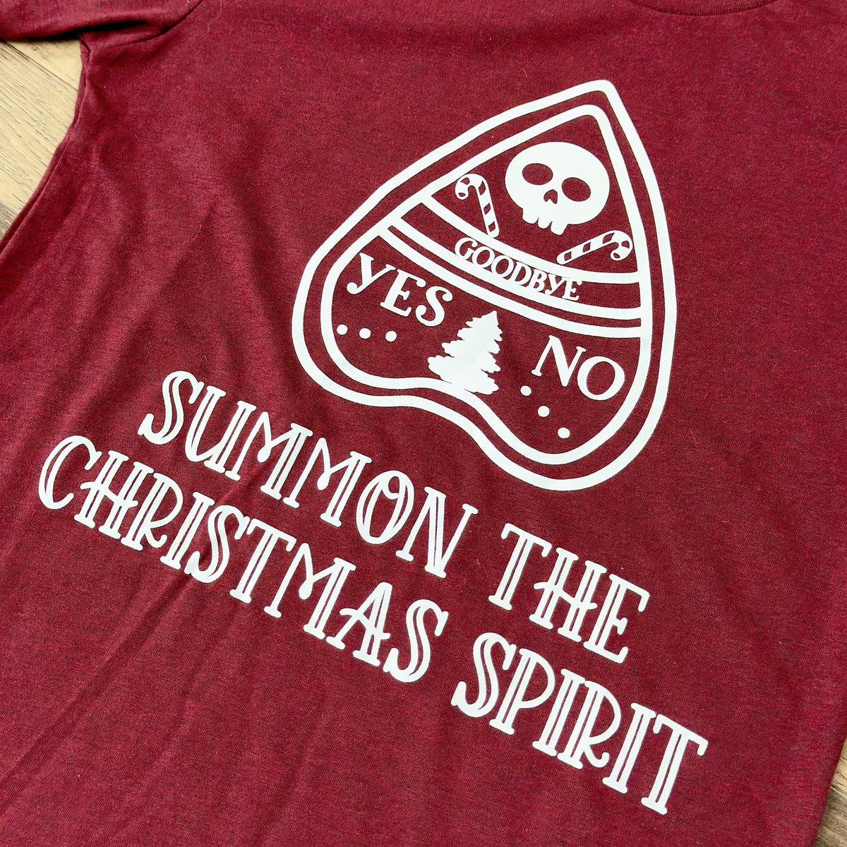The Spellcrafter Summon The Spirit Christmas Tee features a vintage red, textured look with a white Ouija planchette, candy canes, skull, and Christmas tree on soft Heather CVC fabric. Includes "SUMMON THE CHRISTMAS SPIRIT" and Yes/No/Goodbye options.