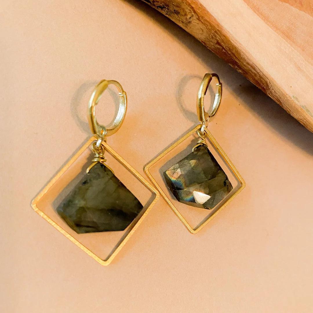 The Spellcrafter Labradorite Framed Diamond Necklace features a faceted dark labradorite stone set in a gold diamond-shaped frame and hangs from hypoallergenic brass wire, shown on a tan surface with a wooden accent.