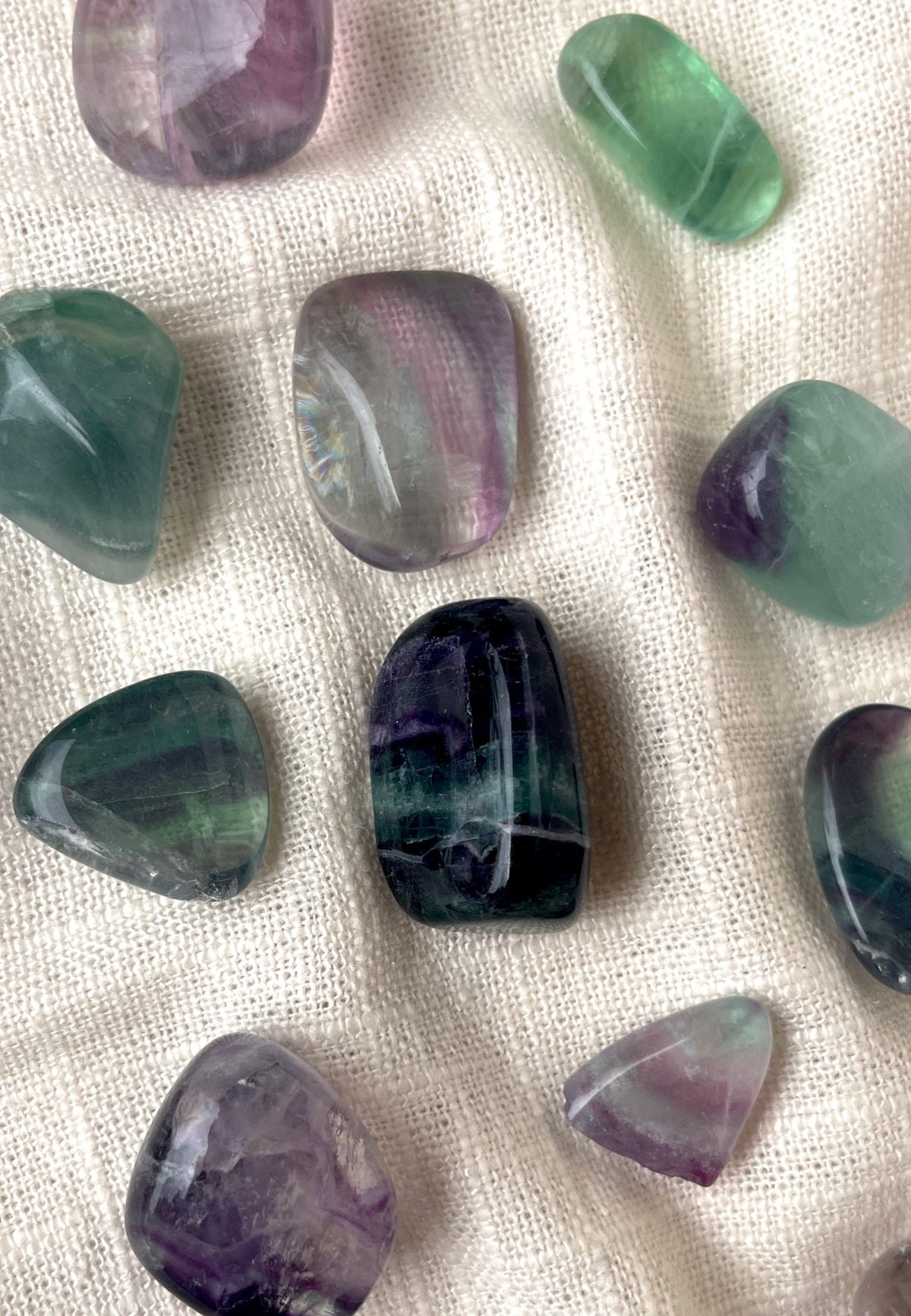 Spellcrafter Fluorite Tumble stones in green, purple, and clear rest on a textured white fabric. Each unique in size and pattern—some with bands—they're prized for grounding and chakra healing.