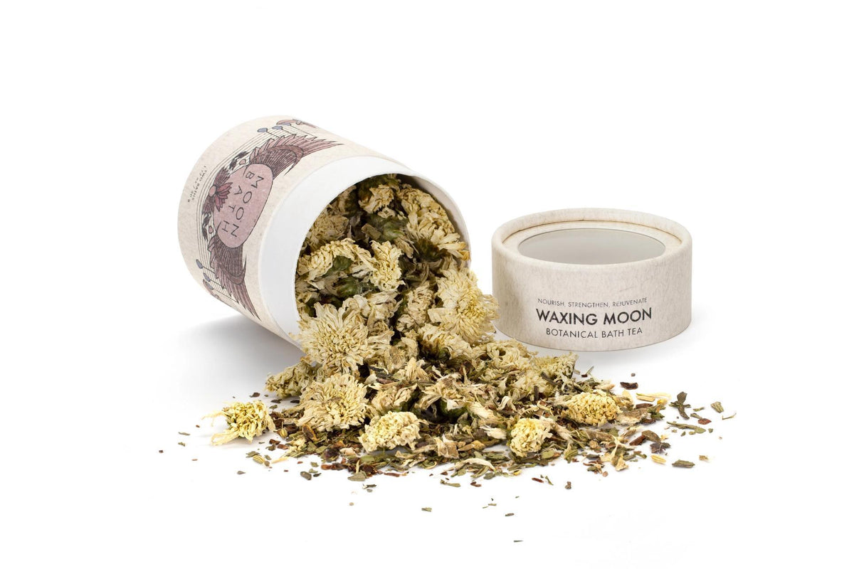 A Spellcrafter Waxing Moon Bath Tea cylindrical container has tipped over, spilling dried flowers and herbs onto a white surface, with its lid resting nearby.
