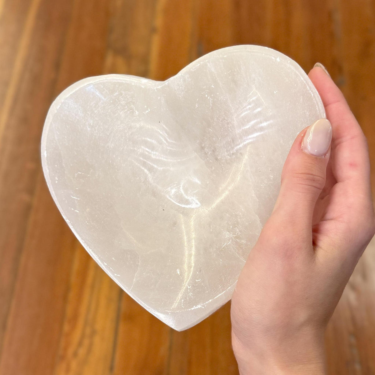 A manicured hand holds the Spellcrafter Selenite Heart Shaped Bowl—valued for its cleansing properties—against wooden floorboards.