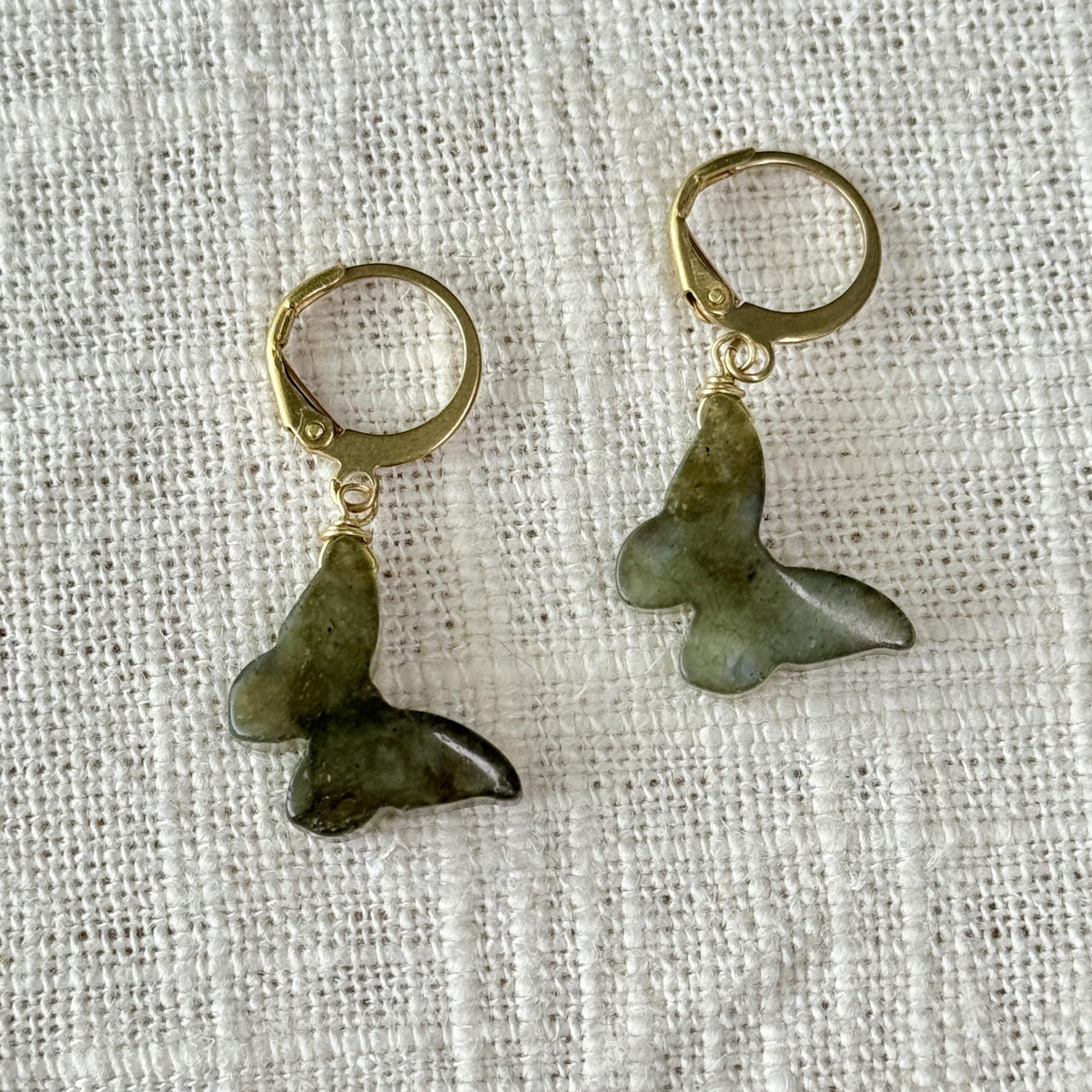 Spellcrafter's Labradorite Butterfly Earrings, featuring gold hoops and green butterfly-shaped labradorite stone pendants, are showcased on a textured white fabric background.