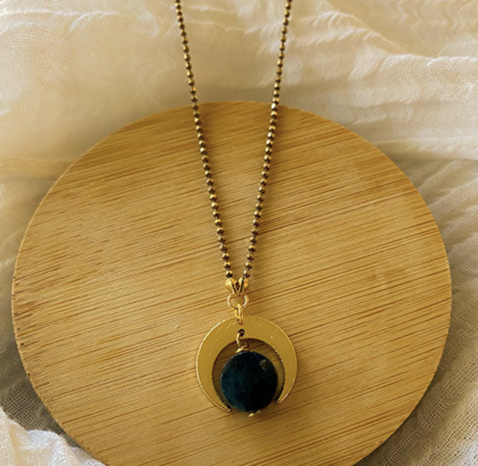 The Spellcrafter Apatite Quarter Moon Necklace showcases a circular blue apatite pendant framed by a brushed brass crescent on a beaded chain, displayed on a round wooden tray with sheer fabric in the background.