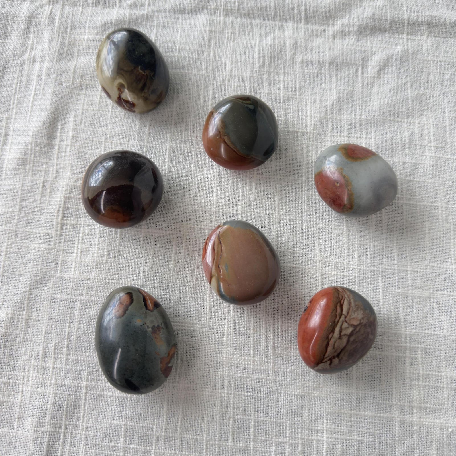 Seven Spellcrafter Polychrome Jasper Palm Stones in brown, gray, red, and cream rest on...