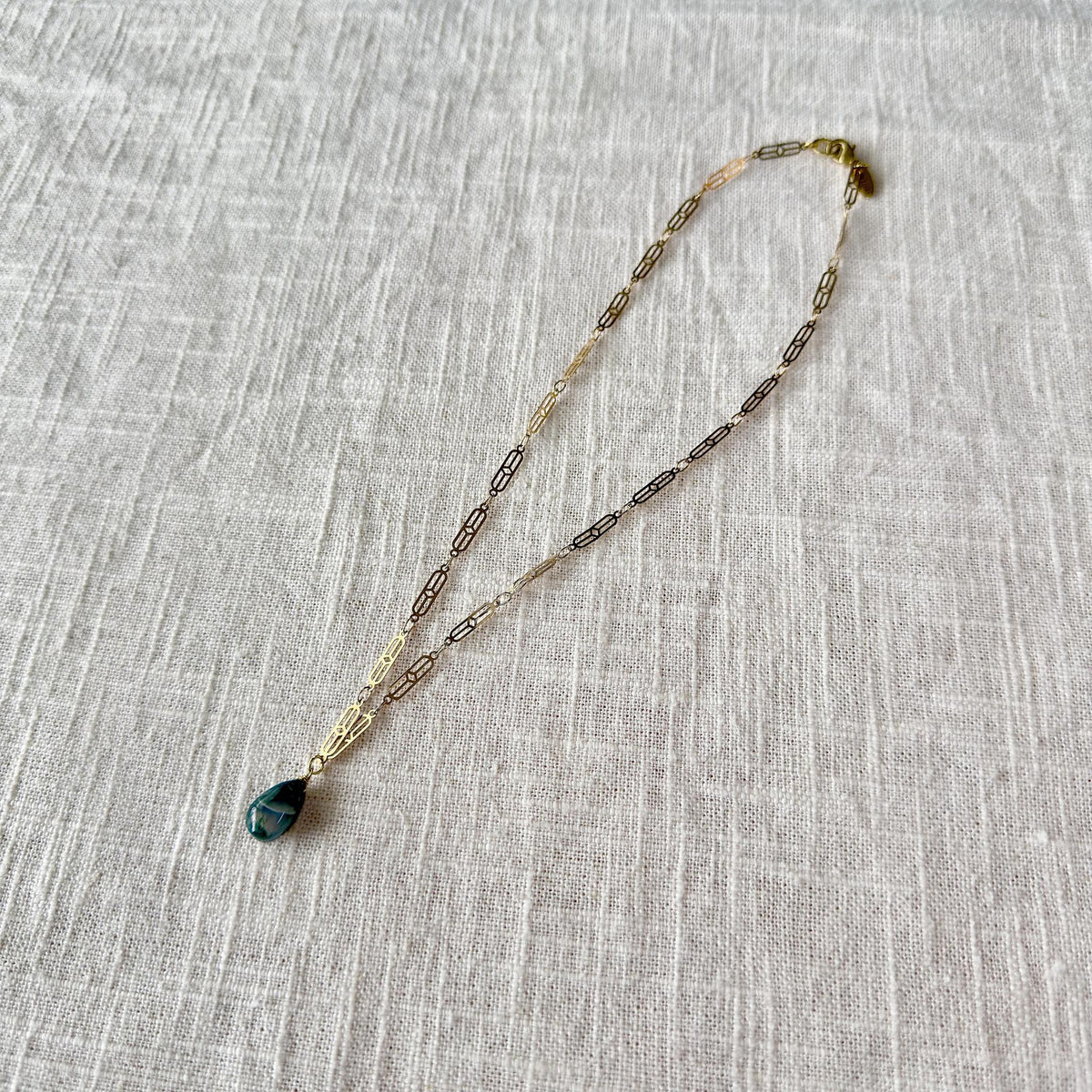 The Spellcrafter Moss Agate Chain Necklace features a gold plated paperclip chain and a teardrop-shaped moss agate pendant, a healing stone, shown on lightly textured fabric.