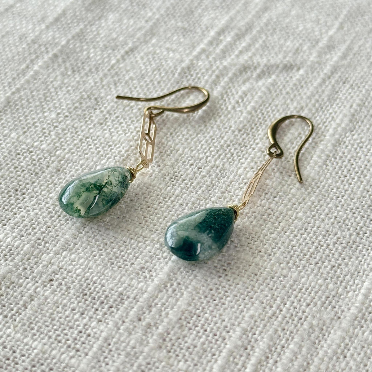 Spellcrafter's Moss Agate Chain Earrings feature green marbled Moss Agate teardrops on gold hooks, elegantly displayed against textured white fabric. Moss Agate is cherished for its gentle healing properties.