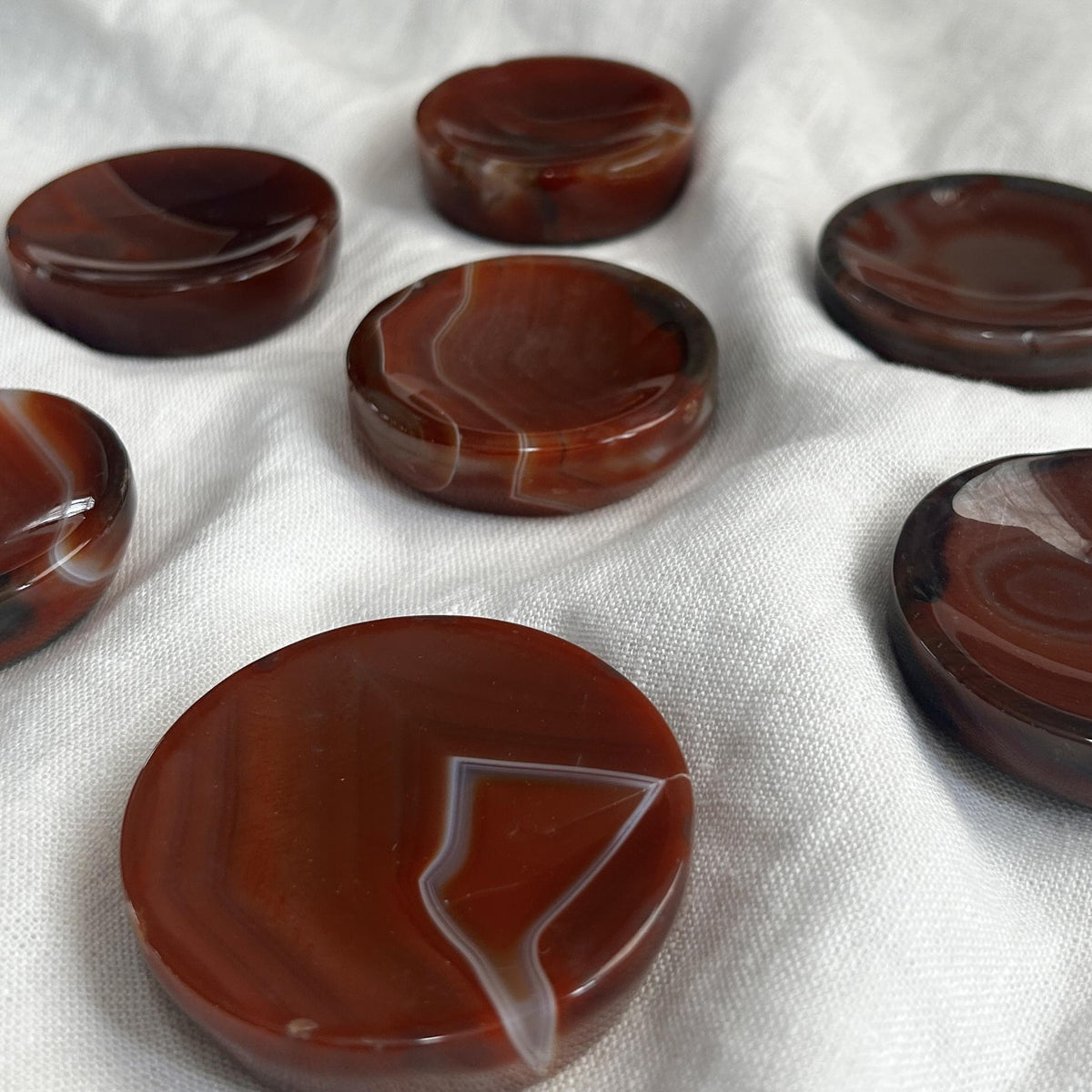 The Spellcrafter Carnelian Crystal Bowl features polished reddish-brown stones with natural white banding, perfect for boosting confidence or balancing chakras. Displayed on a white textured fabric background for added elegance.