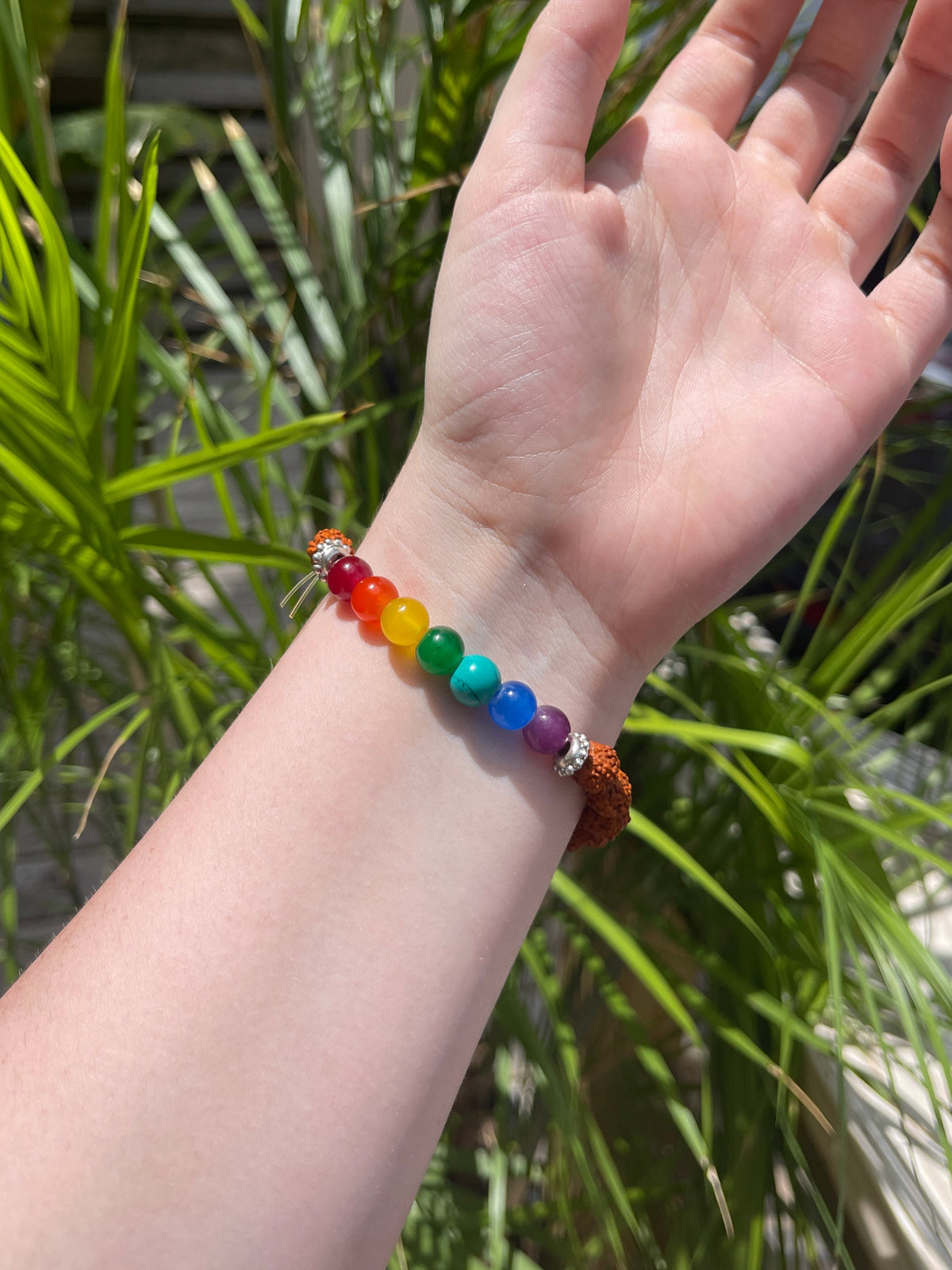 A hand with an outstretched palm wears the Spellcrafter Chakra Stone Bracelet, featuring rainbow-colored beads, with green plants in the background.