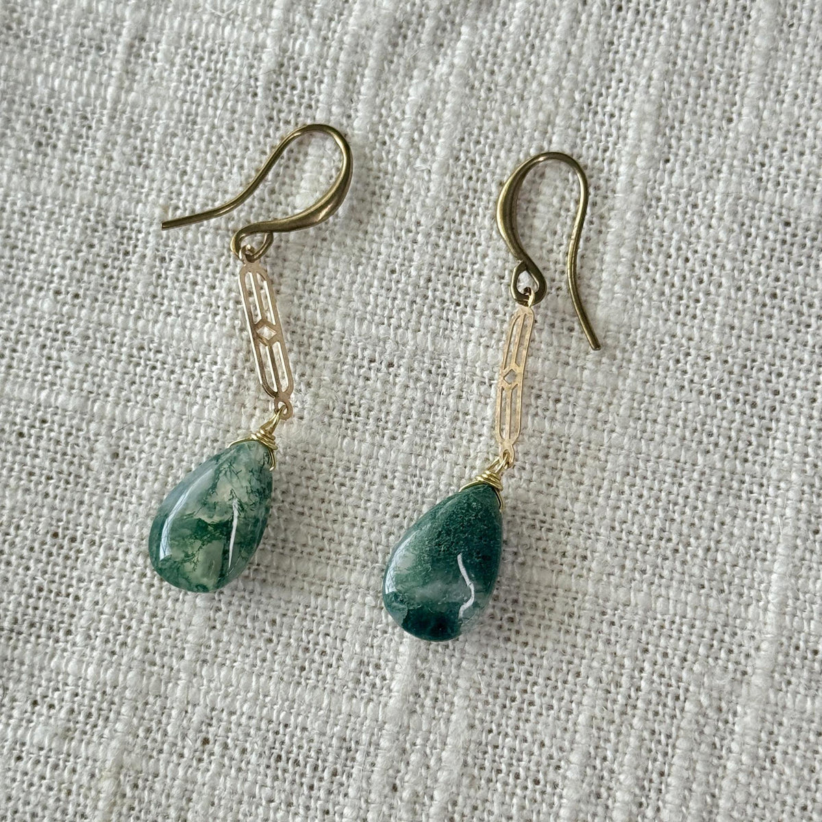 Spellcrafter's Moss Agate Chain Earring showcases teardrop-shaped moss agate with white marbling, hanging from gold hooks and a gold-plated rectangular link chain, displayed on textured white fabric.