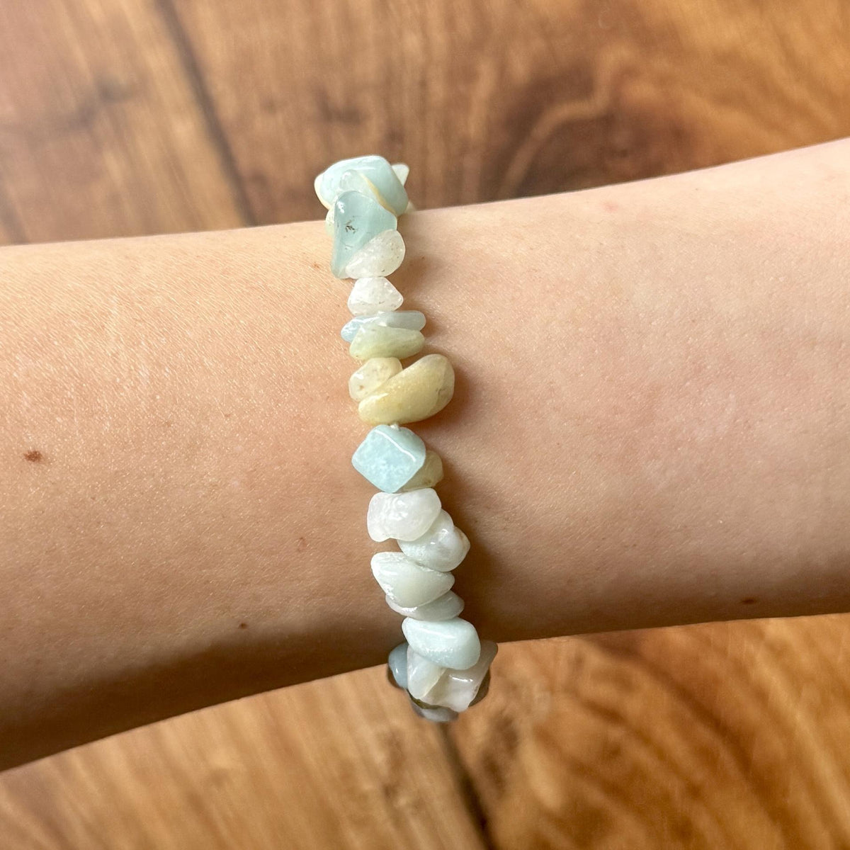 A light-skinned arm wears the Spellcrafter Sunstone Crystal Bracelet, featuring irregular pale blue and off-white gemstone chips on a stretchy band, with a wooden floor visible in the background.