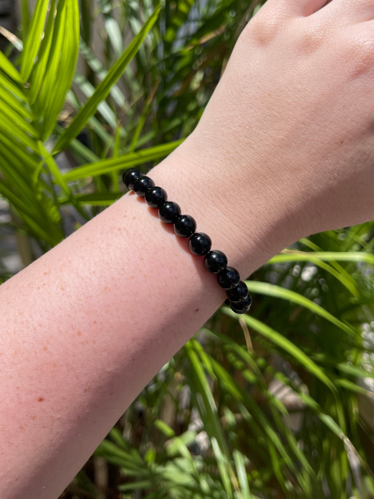 A close-up of a wrist wearing the Spellcrafter Black Sardonyx Bracelet, with green leafy plants in the background.